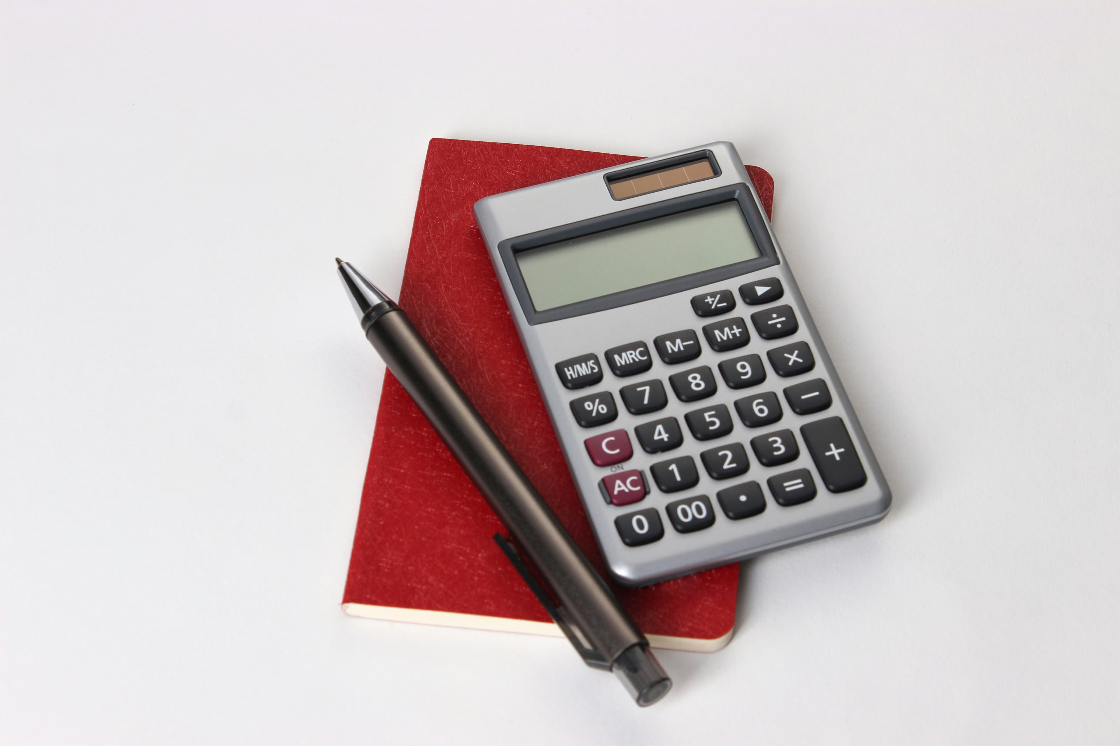 A silver calculator and a dark pen resting on a red notebook against a white background