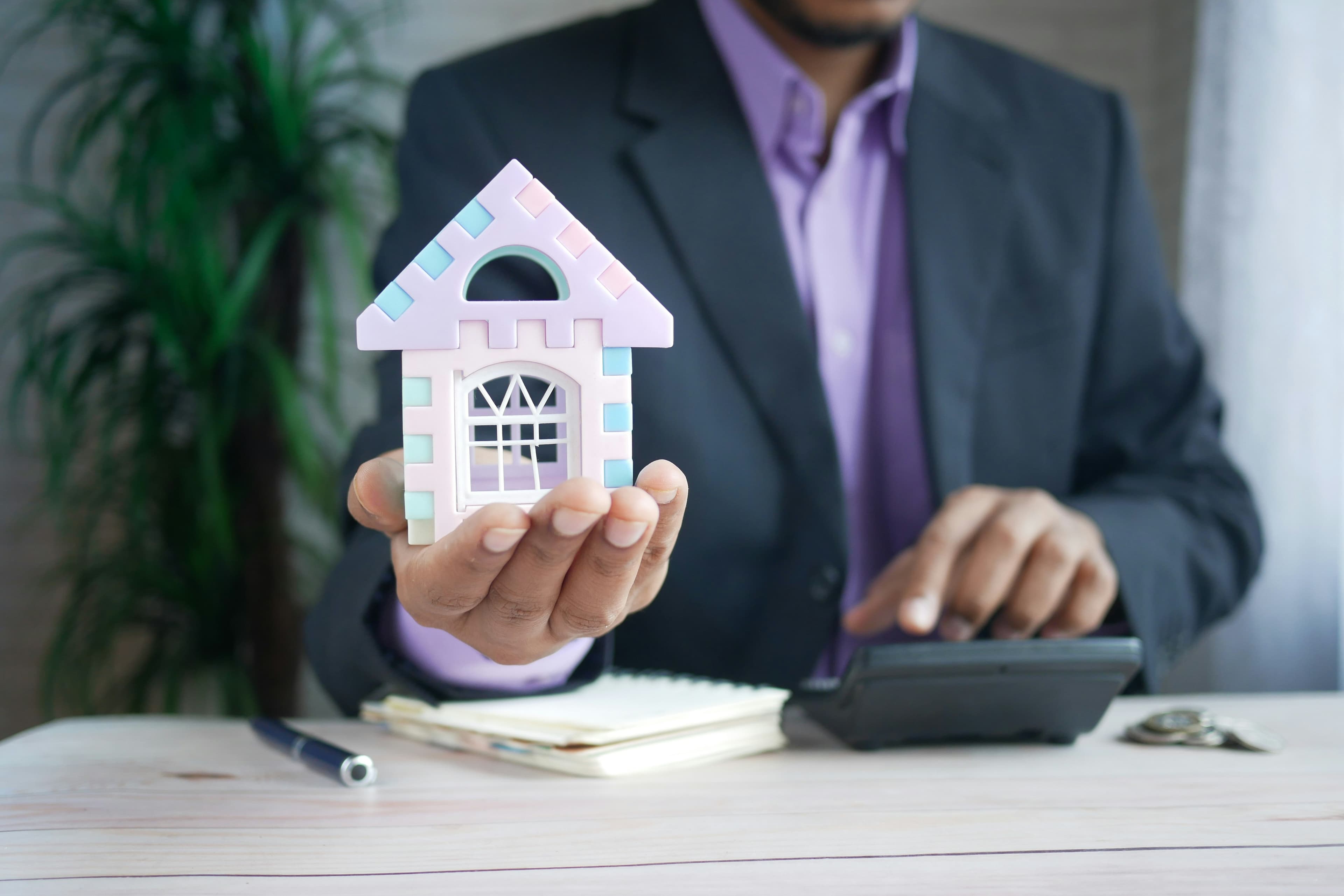 A man at a desk holding a small model house beside a calculator