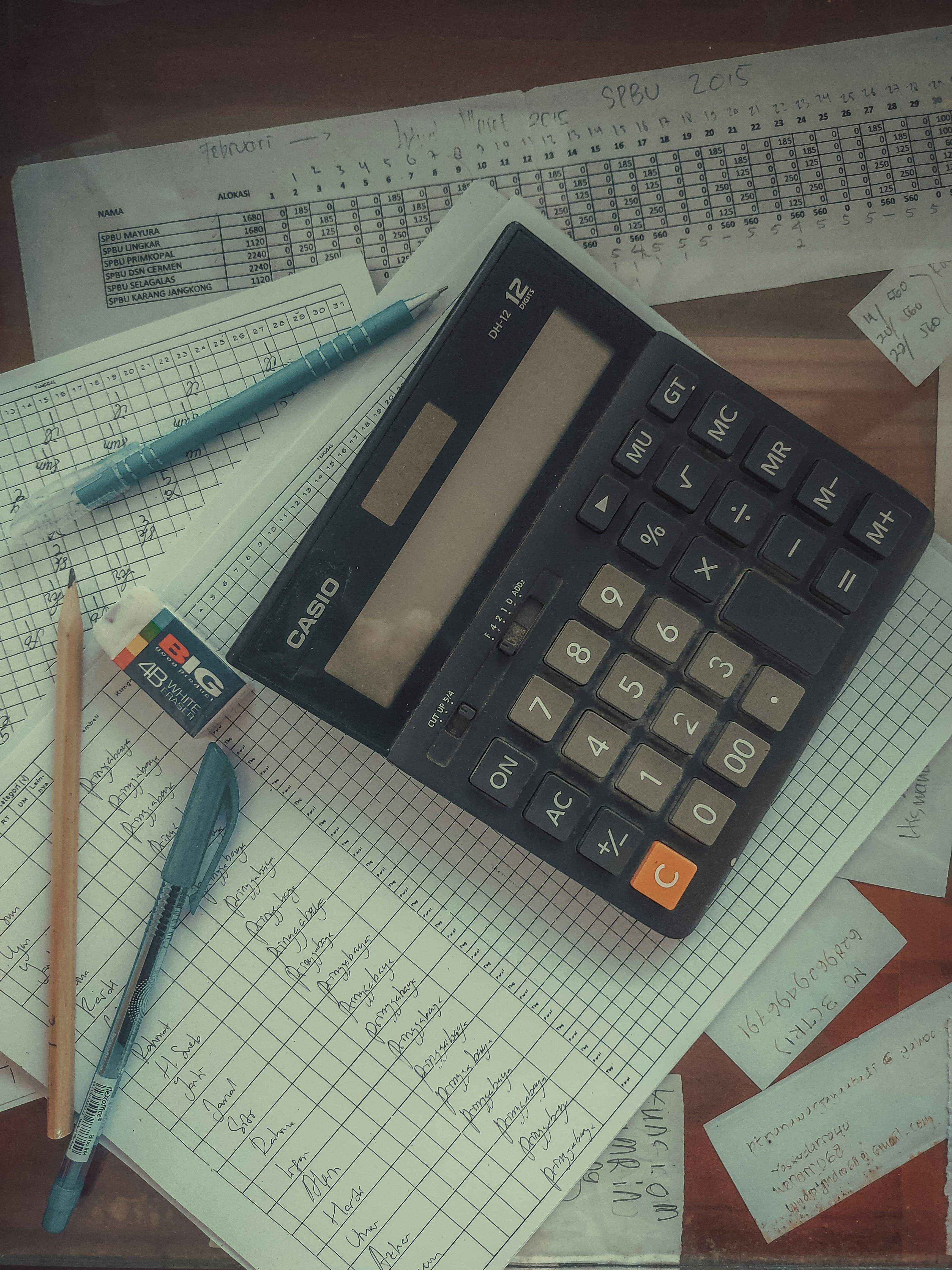 A desk calculator resting on handwritten financial ledgers and pages of figures, with pens alongside.