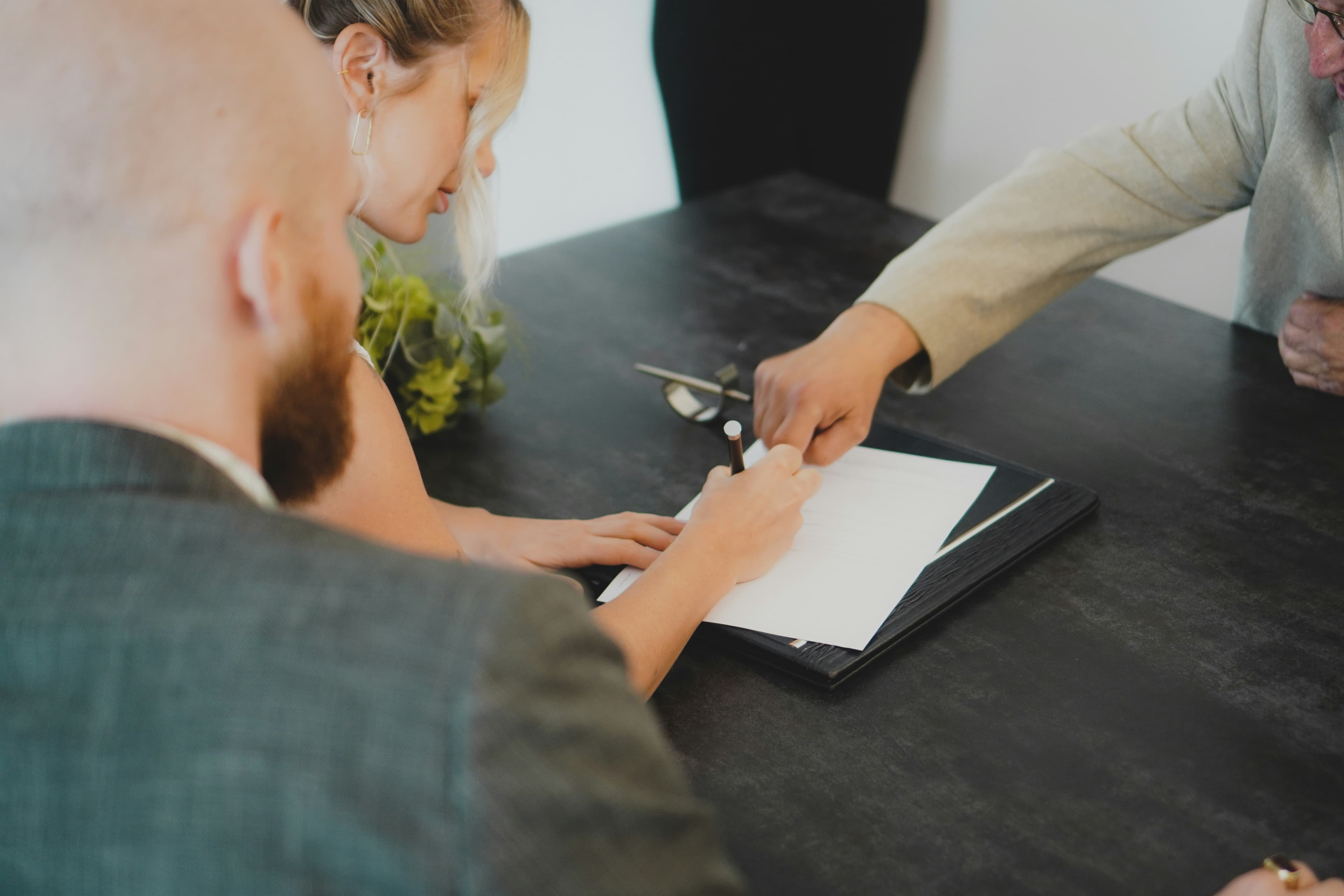 A couple signing a document at a dark desk while another person points to where to sign