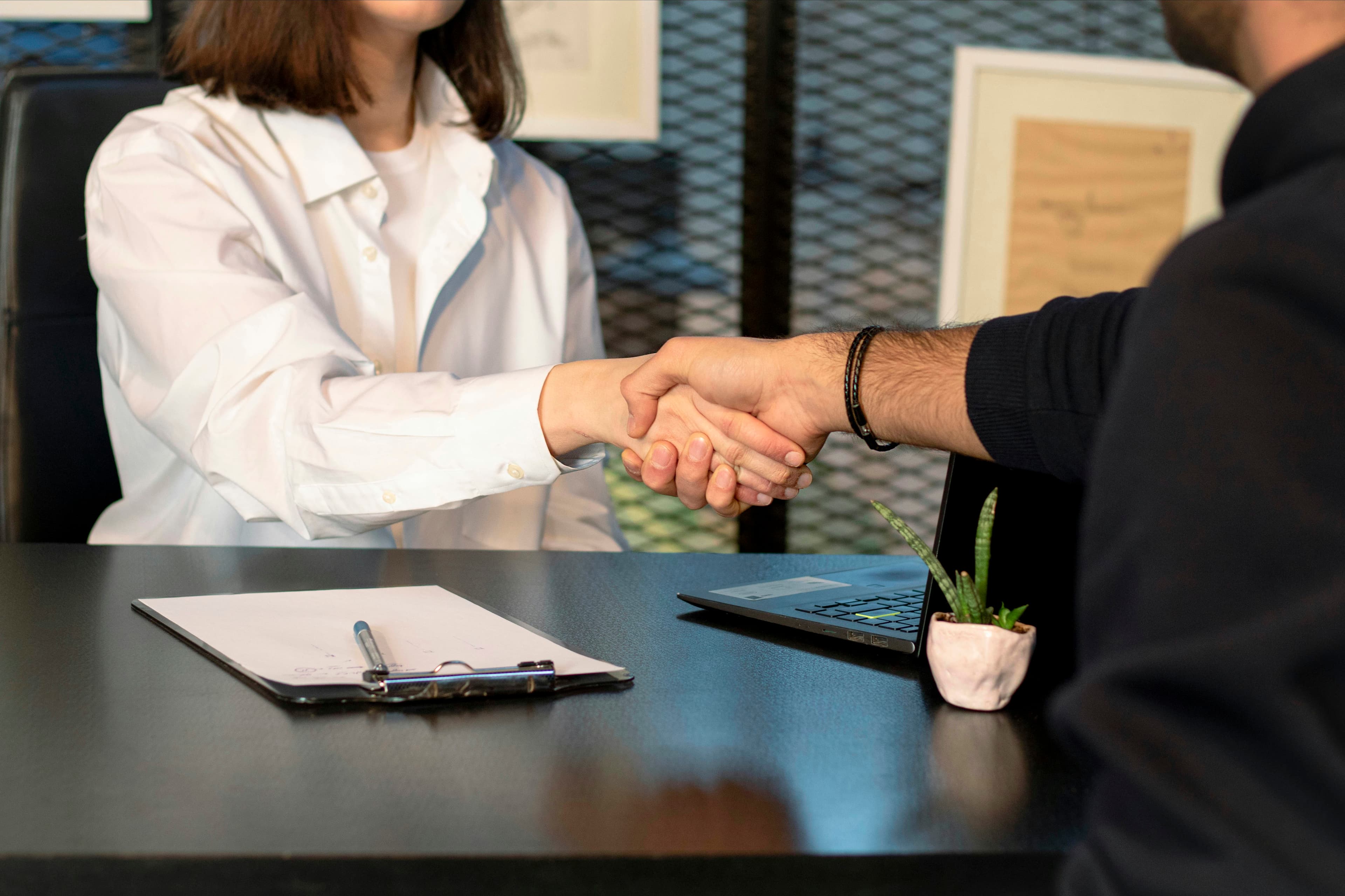 Two people shaking hands across a desk with a clipboard and pen, after agreeing terms
