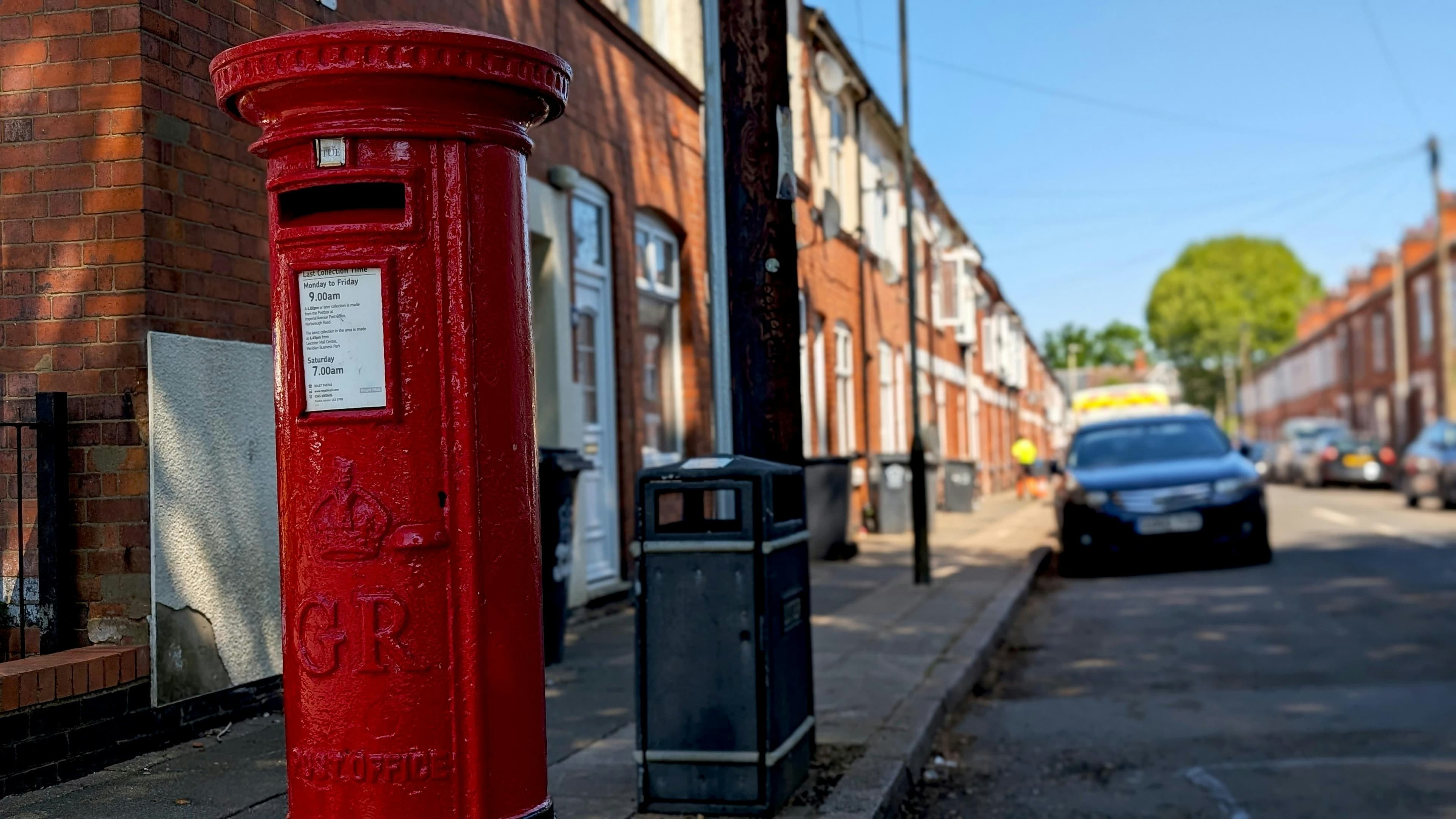 A red Victorian post box on a pavement beside a street of red-brick terraced houses with parked cars