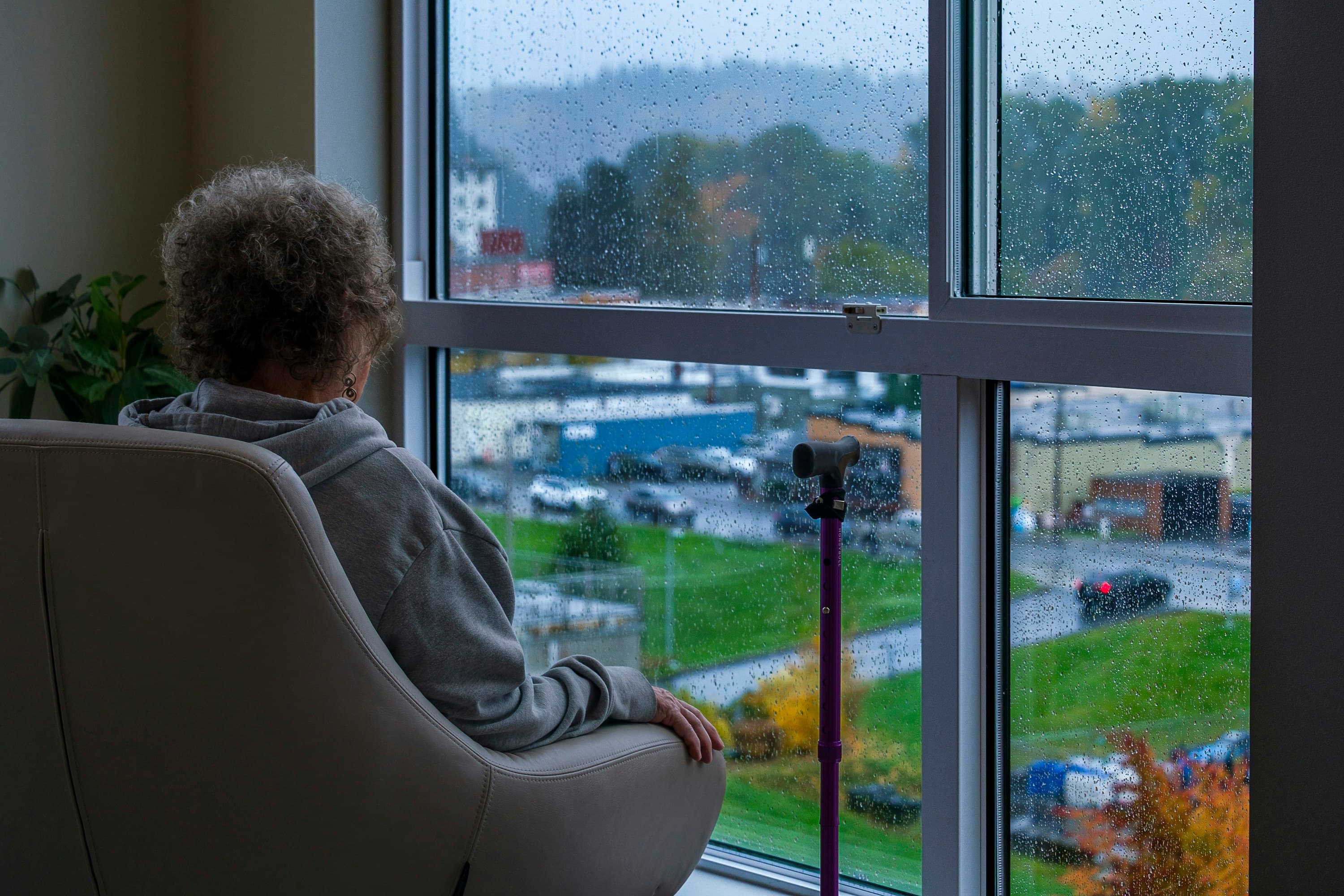 A woman sitting in a chair at home looking out of a rain-streaked window