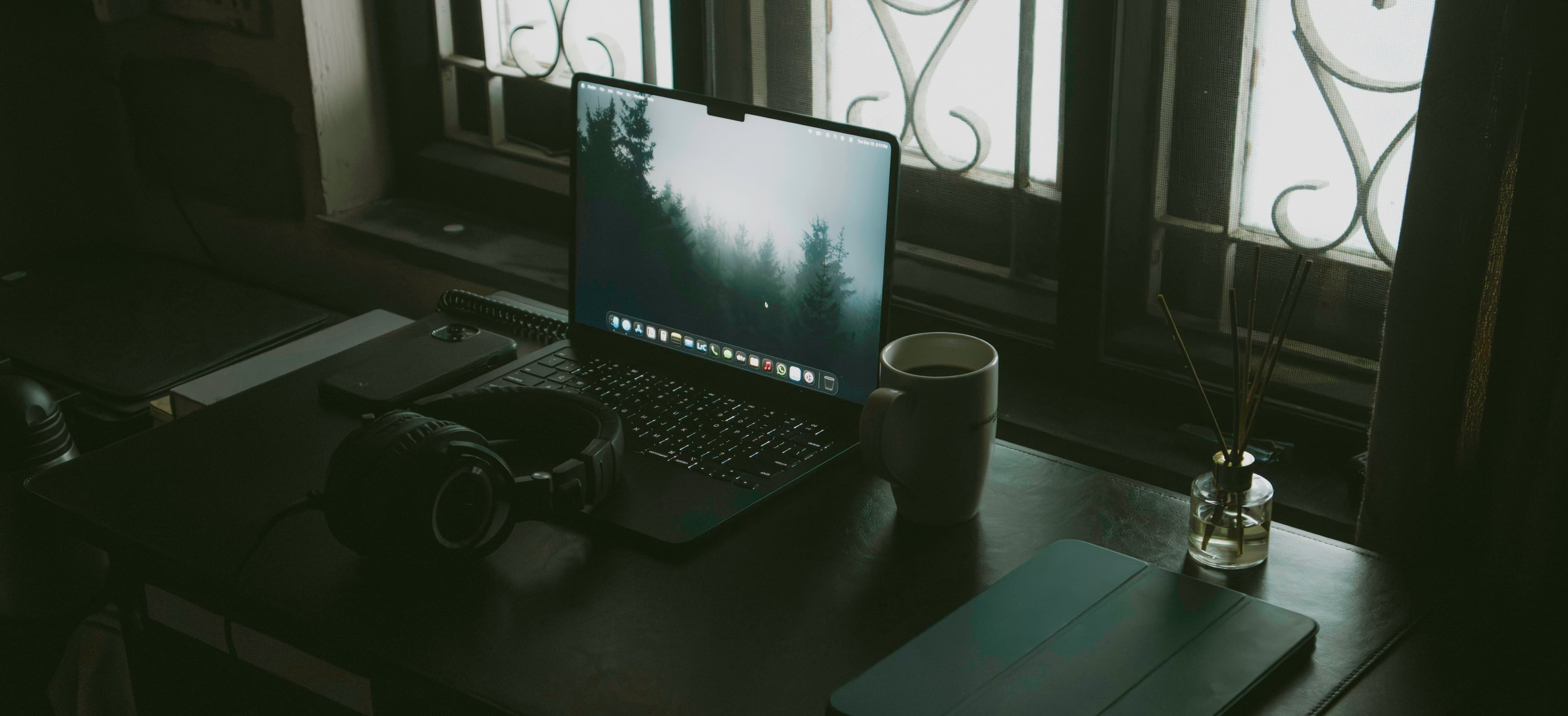 A desk by a window with an open laptop, headphones and a coffee cup