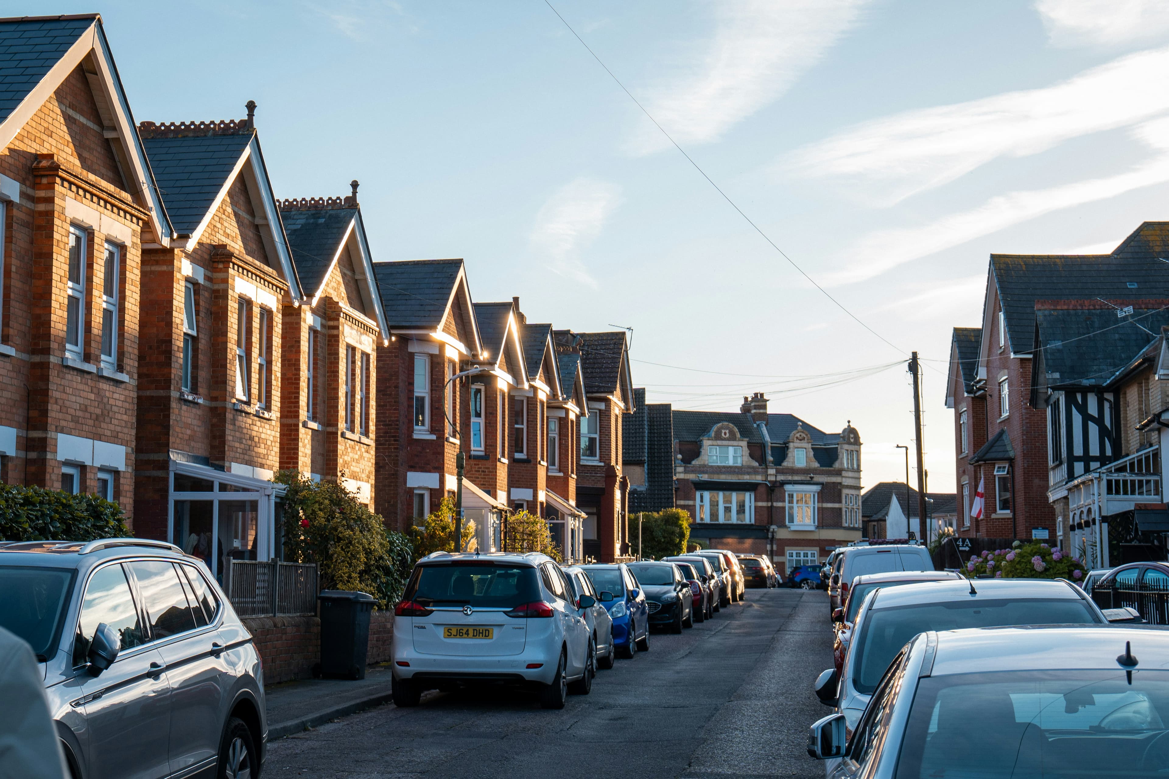 A row of red-brick gabled houses in evening sunlight along a residential street lined with parked cars