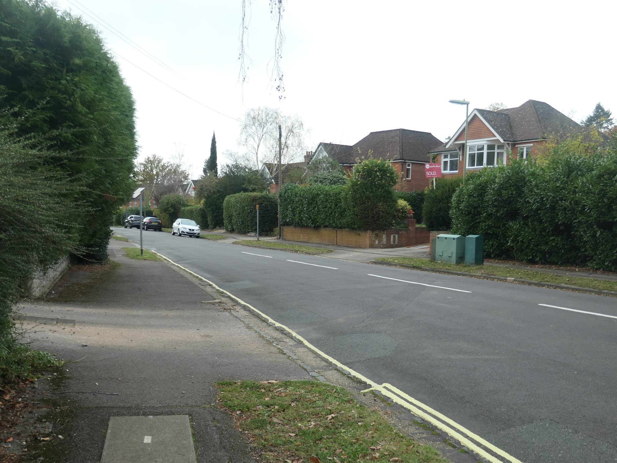 A detached house behind a hedge on a quiet residential road with an estate agent's SOLD board fixed to the front wall