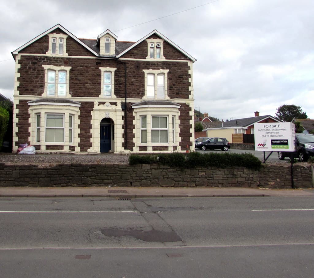 A large stone-built detached house behind a low wall, with an estate agent's For Sale board on the roadside verge