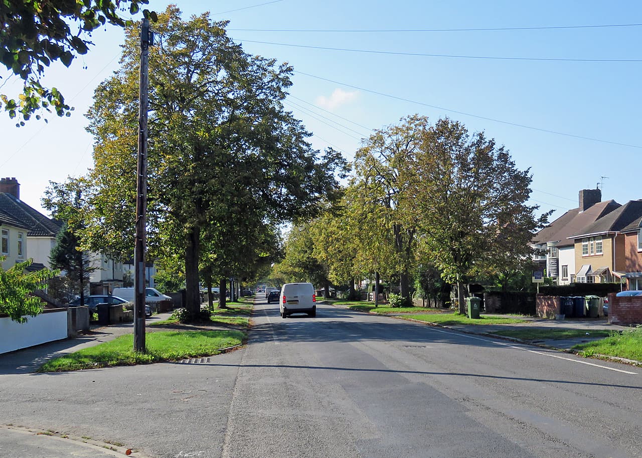 A quiet tree-lined suburban road in early autumn, with parked cars, a van driving away and an estate agent's board outside a house on the right