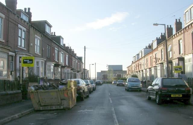 A street of red brick terraced houses with estate agents' For Sale boards on both sides of the road under a clear winter sky