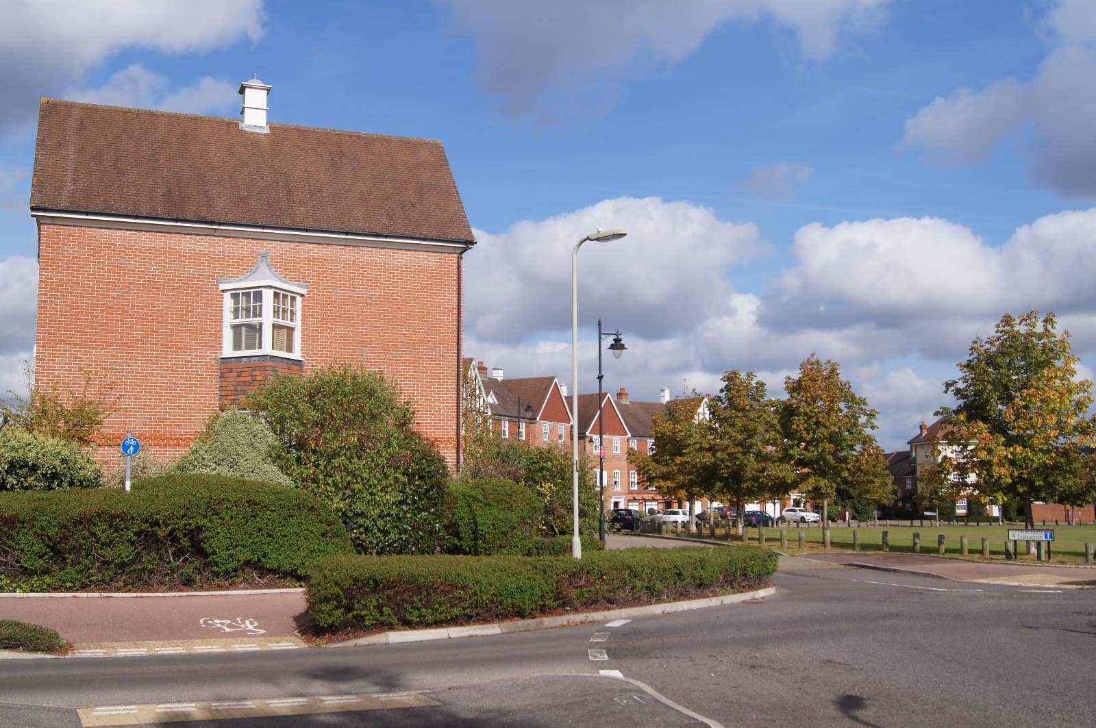 Modern red brick houses around a green on a quiet crescent, with a street lamp, trimmed hedges and autumn trees under a blue sky
