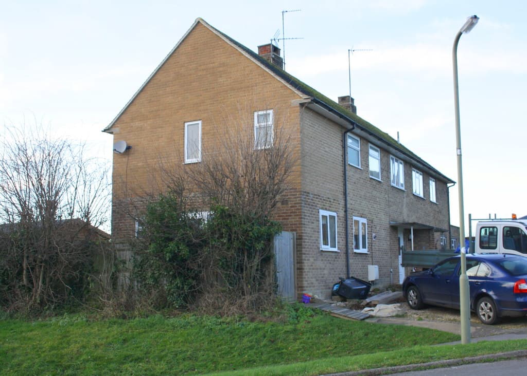 A pair of brick semi-detached houses on a grass bank with an overgrown front garden and a car parked beside them under a pale sky