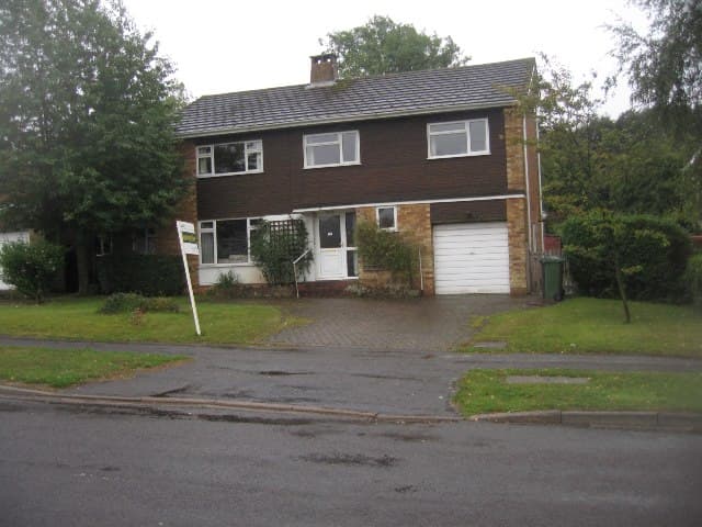 A detached house set back behind a lawn on a damp day, with an estate agent's board leaning at the edge of the block paved drive