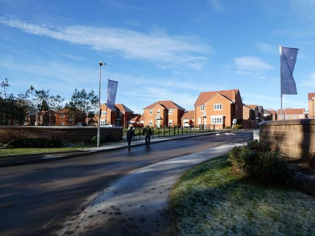 A newly built housing estate of brick homes on the edge of a village, with parked cars and young front gardens