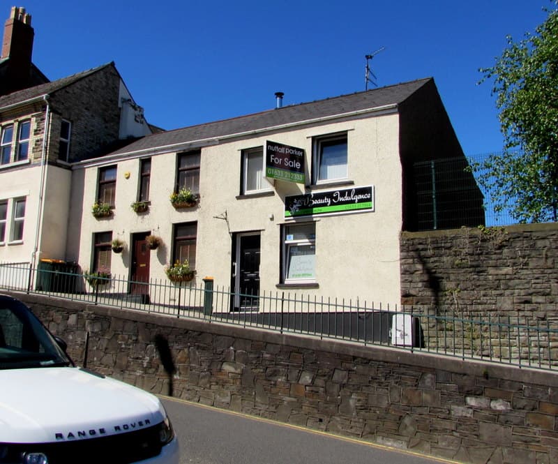 A For Sale board fixed to the wall of a rendered house on a sunny street corner above a stone retaining wall
