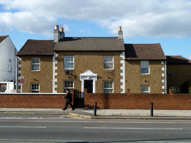 A large house for sale set back behind a low brick wall on a main road, with an estate agent's board at the roadside and a passer-by walking past the gate