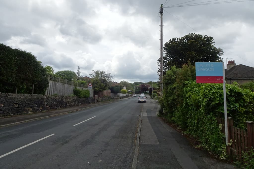 An estate agent's Sold board on a post beside a hedge on a quiet residential road under grey cloud