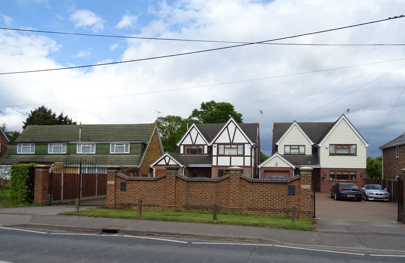 Two large modern detached houses with mock Tudor and weatherboard fronts behind a brick wall on Noak Hill Road in Billericay, with a smaller semi-detached house beside them