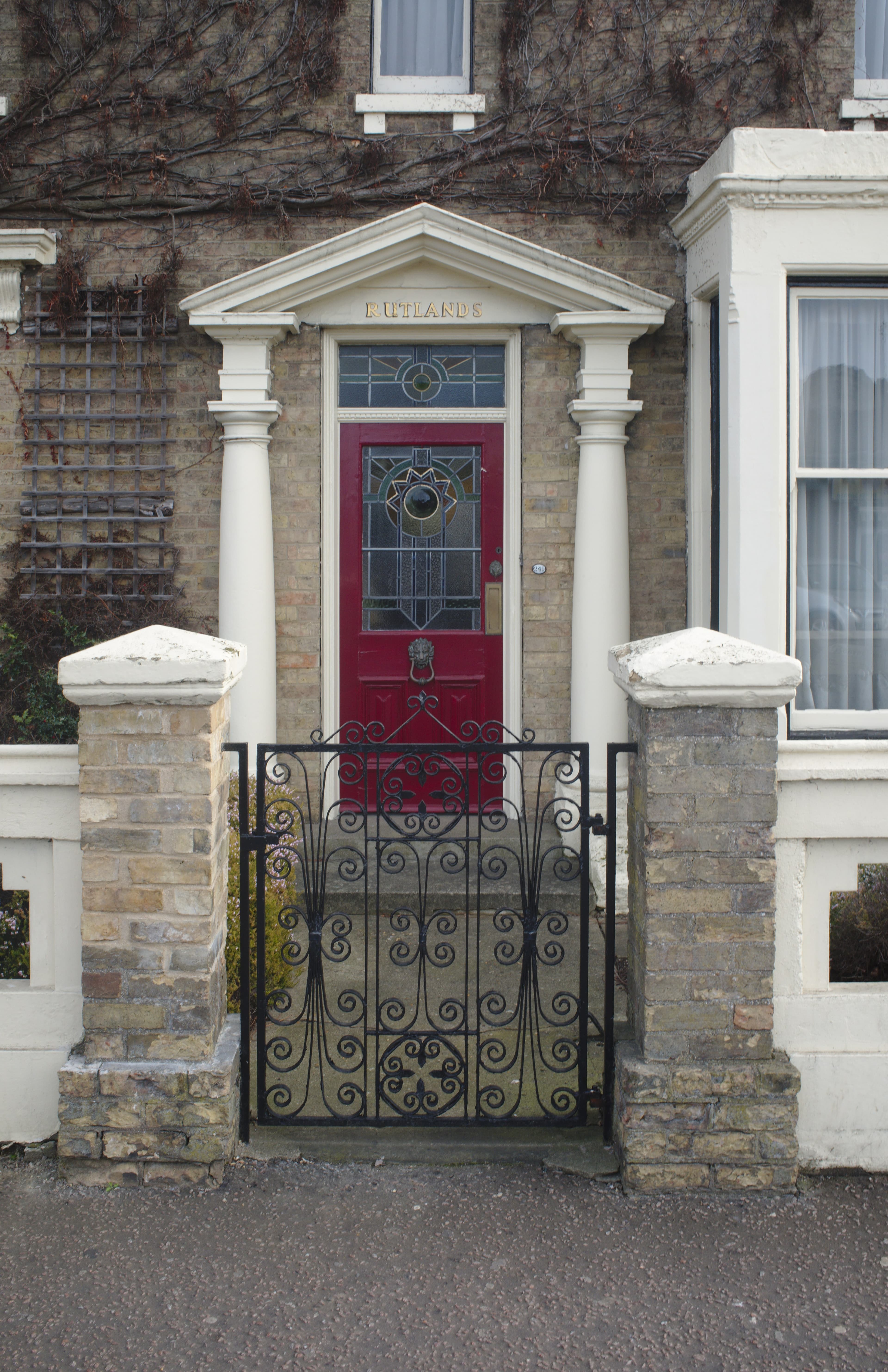 A red front door with a stained-glass panel under a white pillared porch, behind a black wrought-iron gate between stone pillars