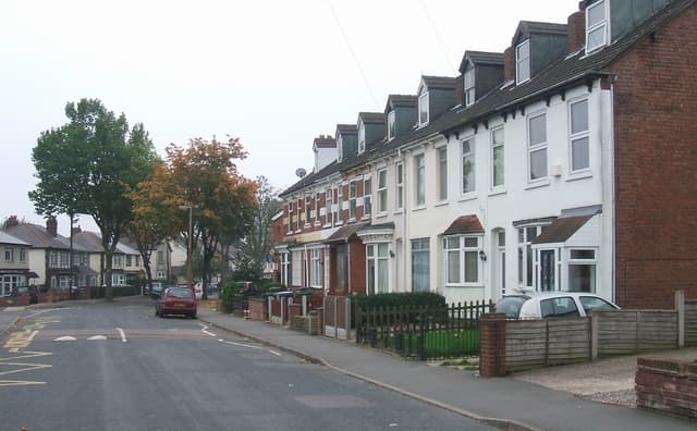 A quiet residential road of bay-fronted terraced houses on an overcast autumn day, with turning leaves on the roadside trees
