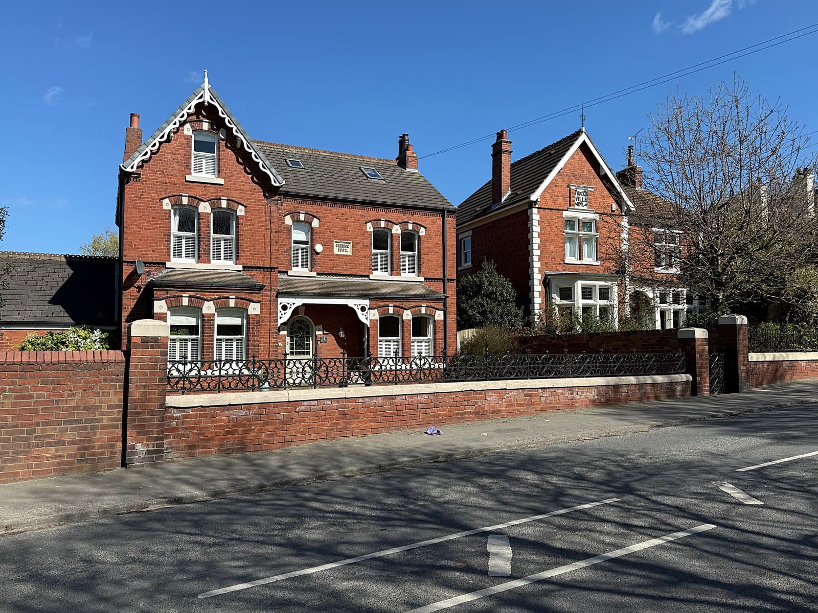 Two large red brick detached Victorian houses behind a low brick garden wall on a quiet residential road under a clear blue sky