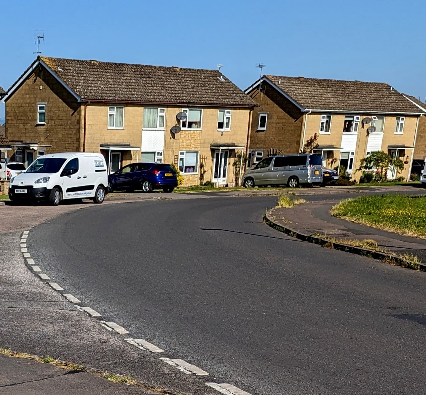 A quiet residential cul-de-sac of stone-faced semi-detached houses with cars parked on the drives under a clear blue sky