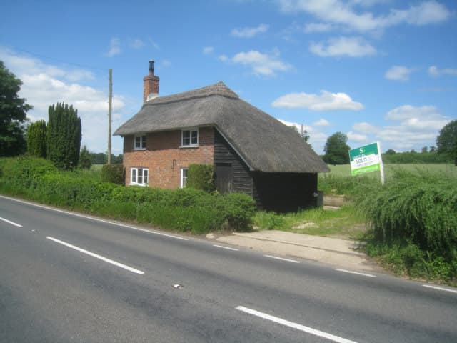 A thatched brick cottage beside a country road in summer, with an estate agent's Sold board standing in the hedge alongside