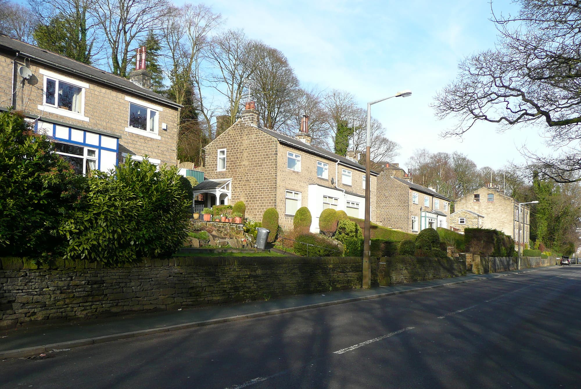 A row of stone semi-detached houses behind a dry-stone wall and clipped hedges on a sloping residential road under a clear blue sky