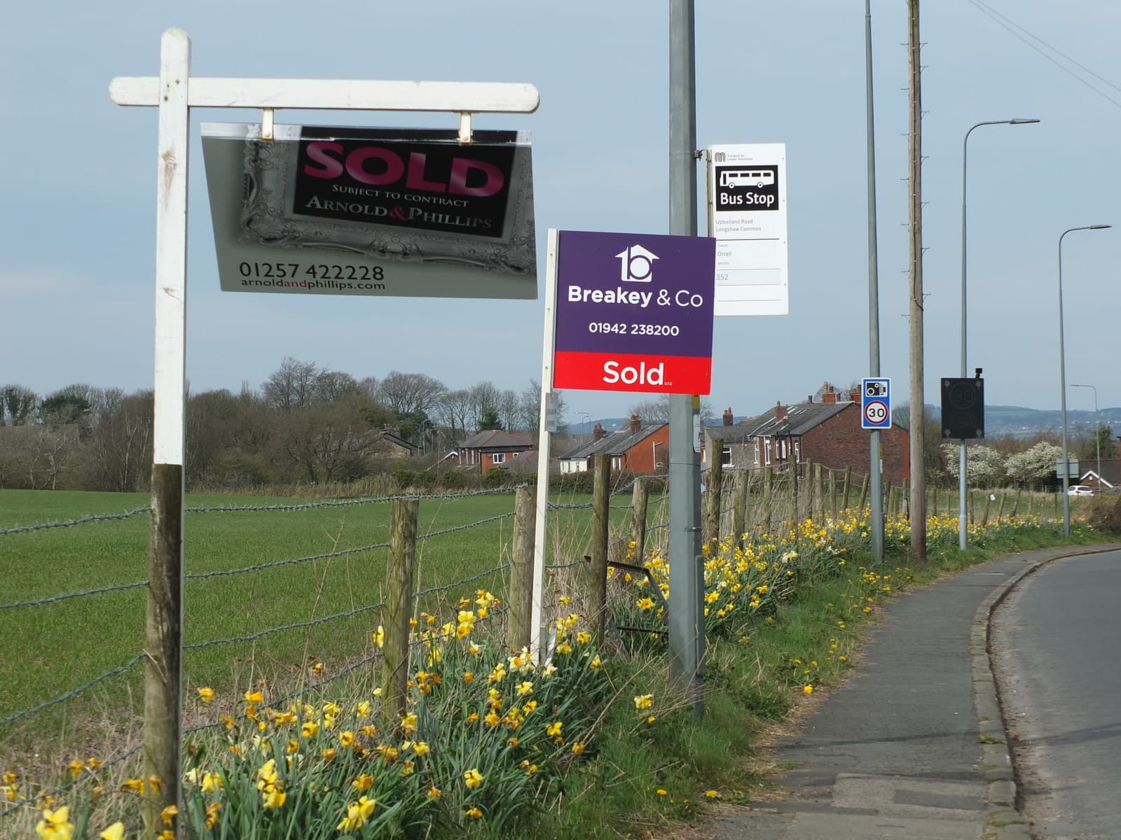 Two estate agent For Sale signs outside houses on a residential road in Billinge
