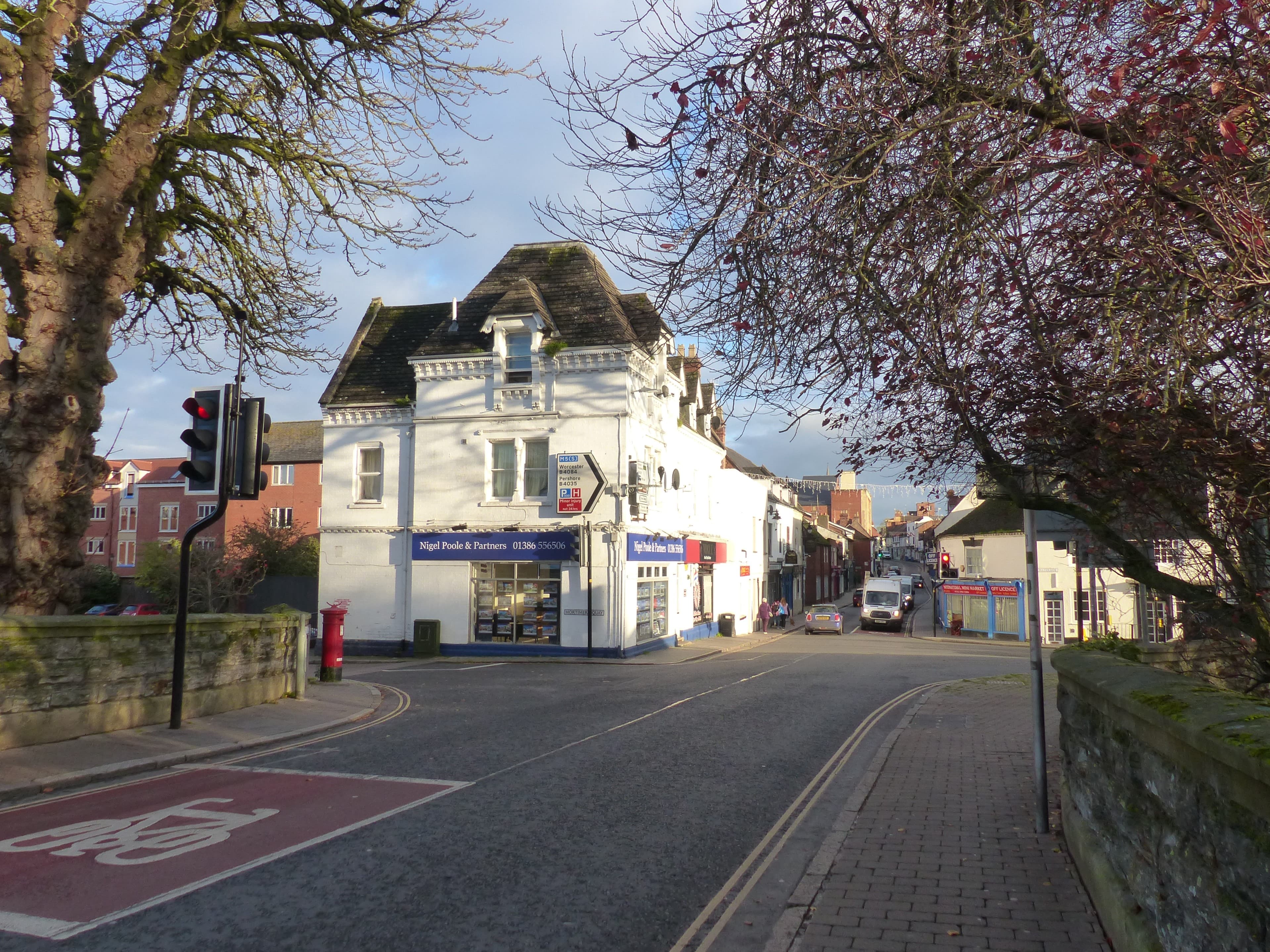 A white corner building housing an estate agent's office at a quiet town road junction, with traffic lights and bare trees in low evening sun