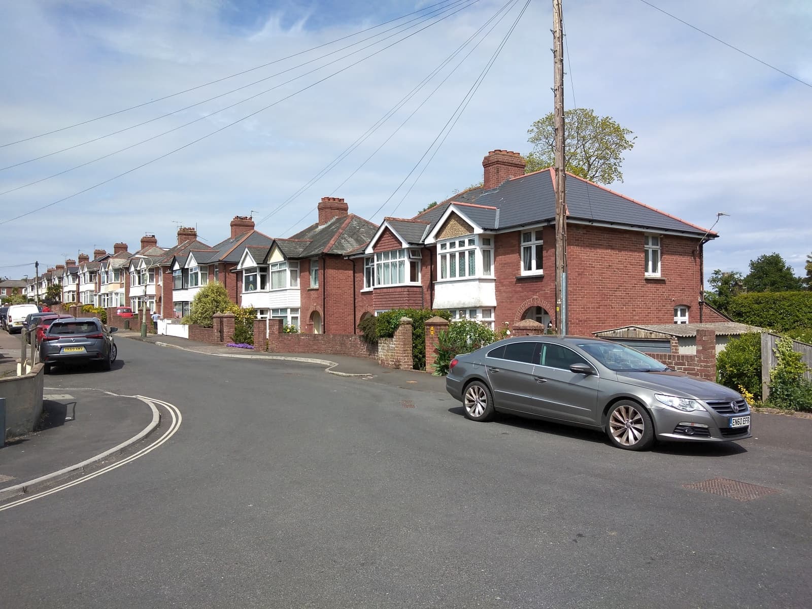 A curving residential avenue of red-brick semi-detached houses with bay windows in Heavitree, Exeter, with cars parked on the street