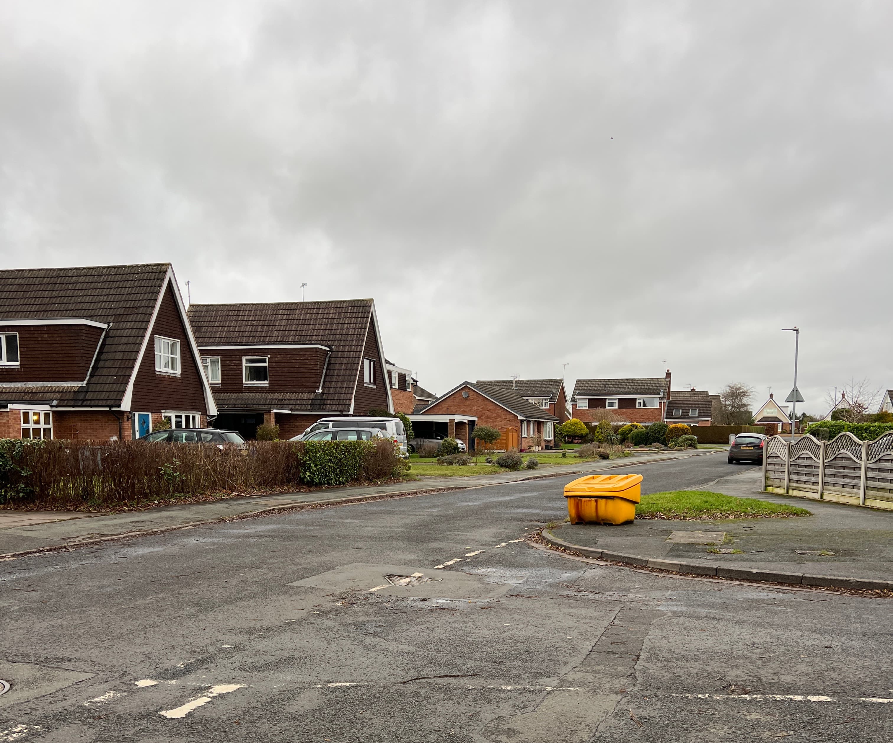 A quiet residential street of brick and tile-hung houses with cars on the drives, seen from a junction on a grey overcast day