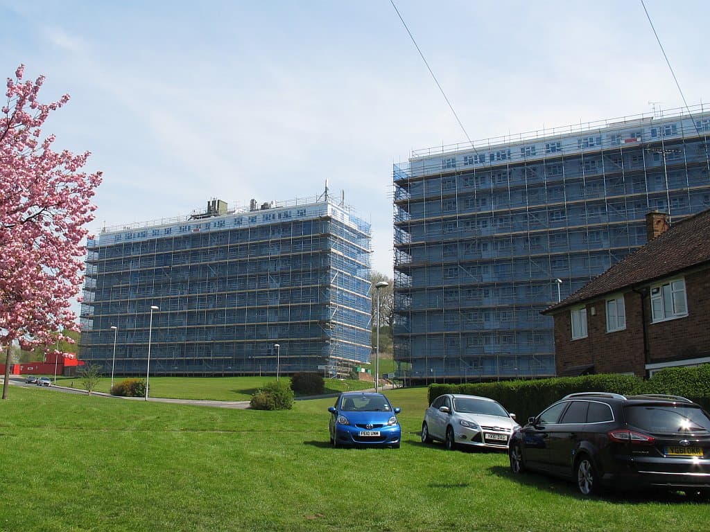 Two blocks of flats wrapped in scaffolding and protective sheeting during refurbishment, seen across a grass bank with a blossom tree