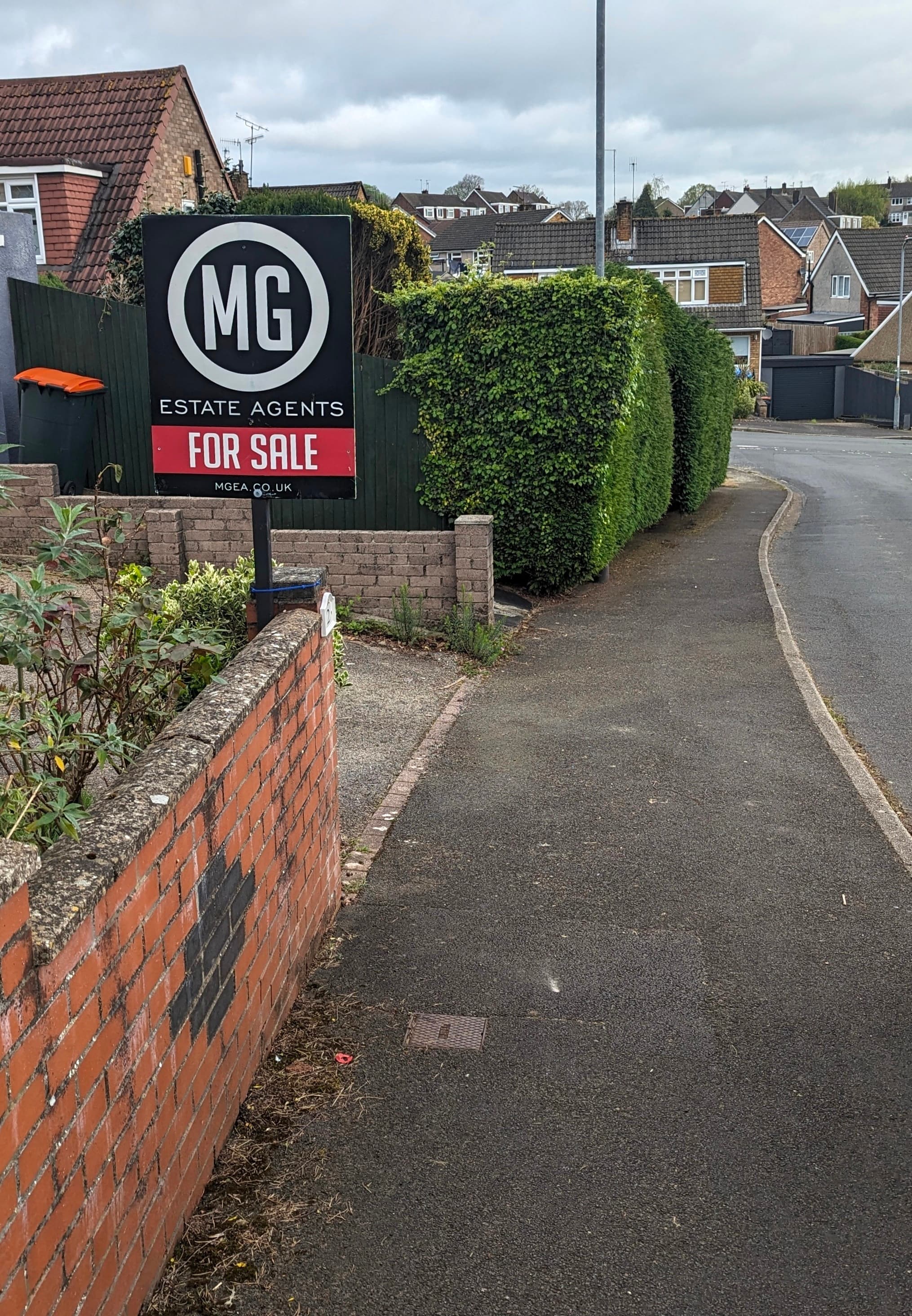 An estate agent's For Sale board on a post beside a brick garden wall, with a suburban street of houses curving away behind it