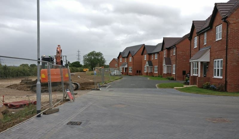 A finished street of red brick new build houses beside a fenced construction area with a digger, on a grey day
