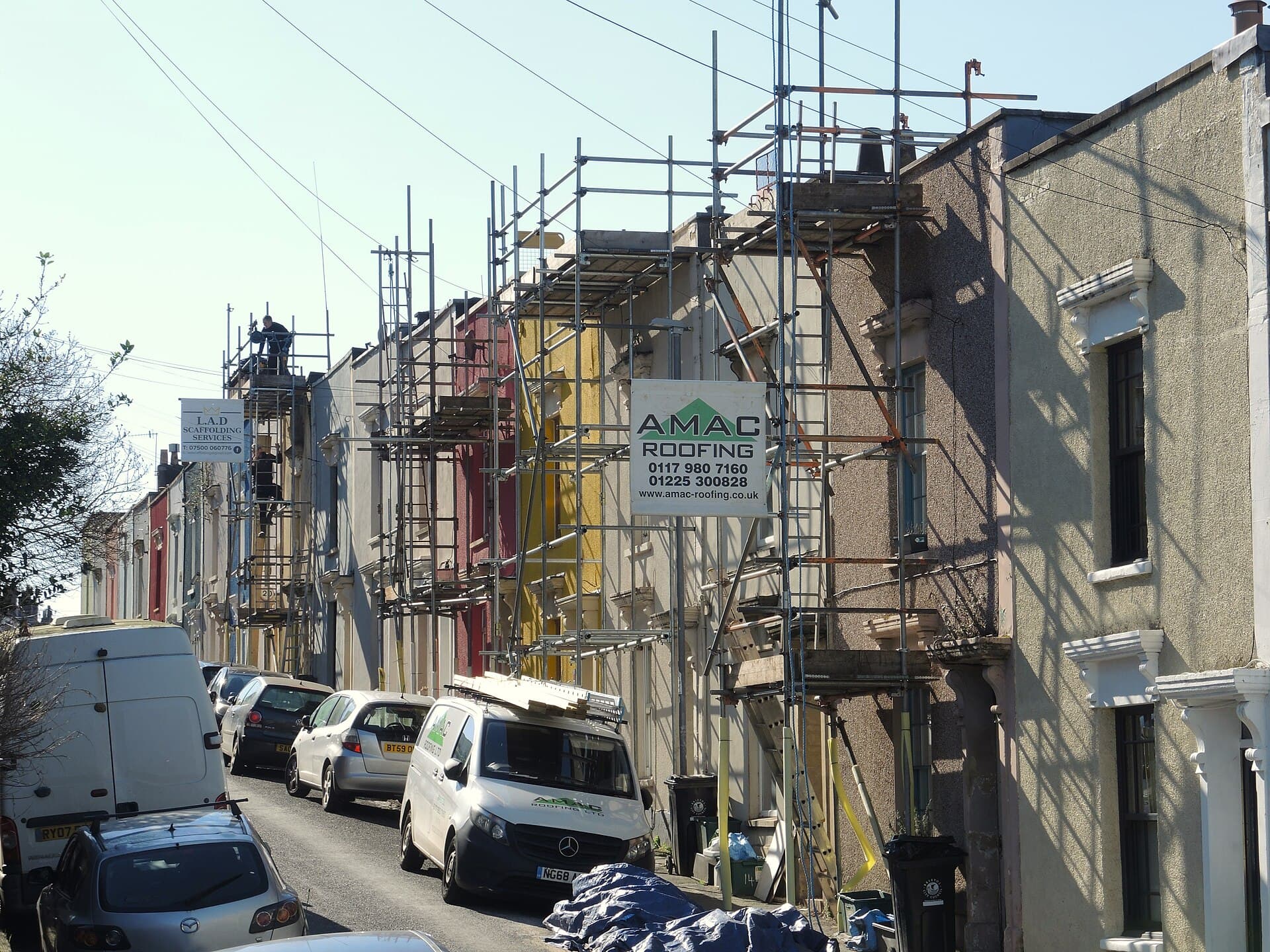 Scaffolding erected along a terrace of painted houses with a roofing contractor's van parked outside and workers on the scaffold boards