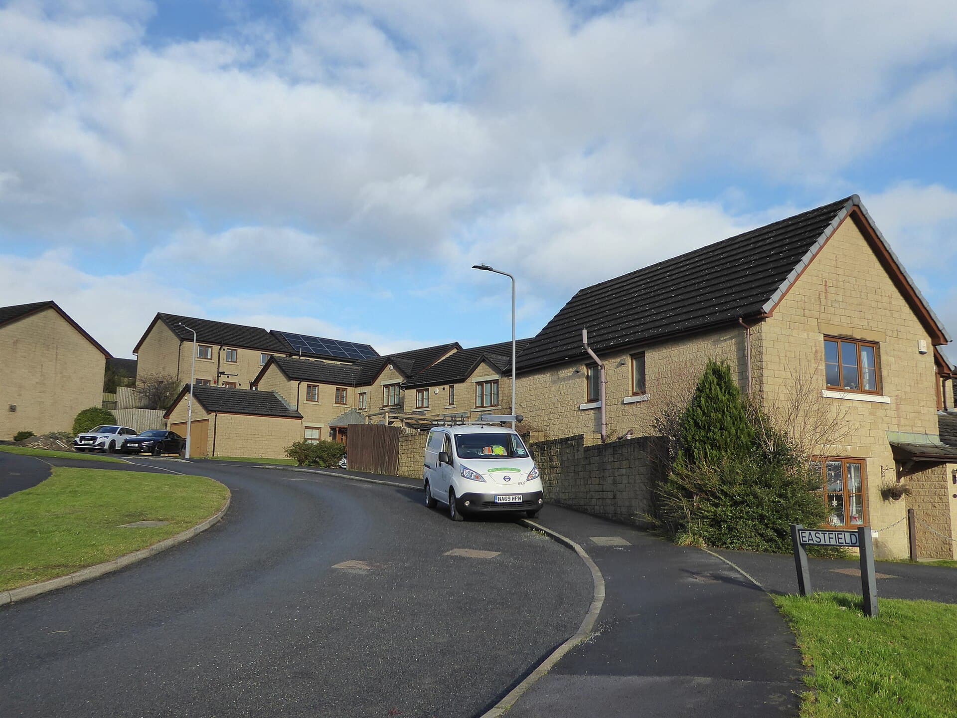 A curving road through a modern estate of stone-built detached and semi-detached houses under a bright, cloudy sky
