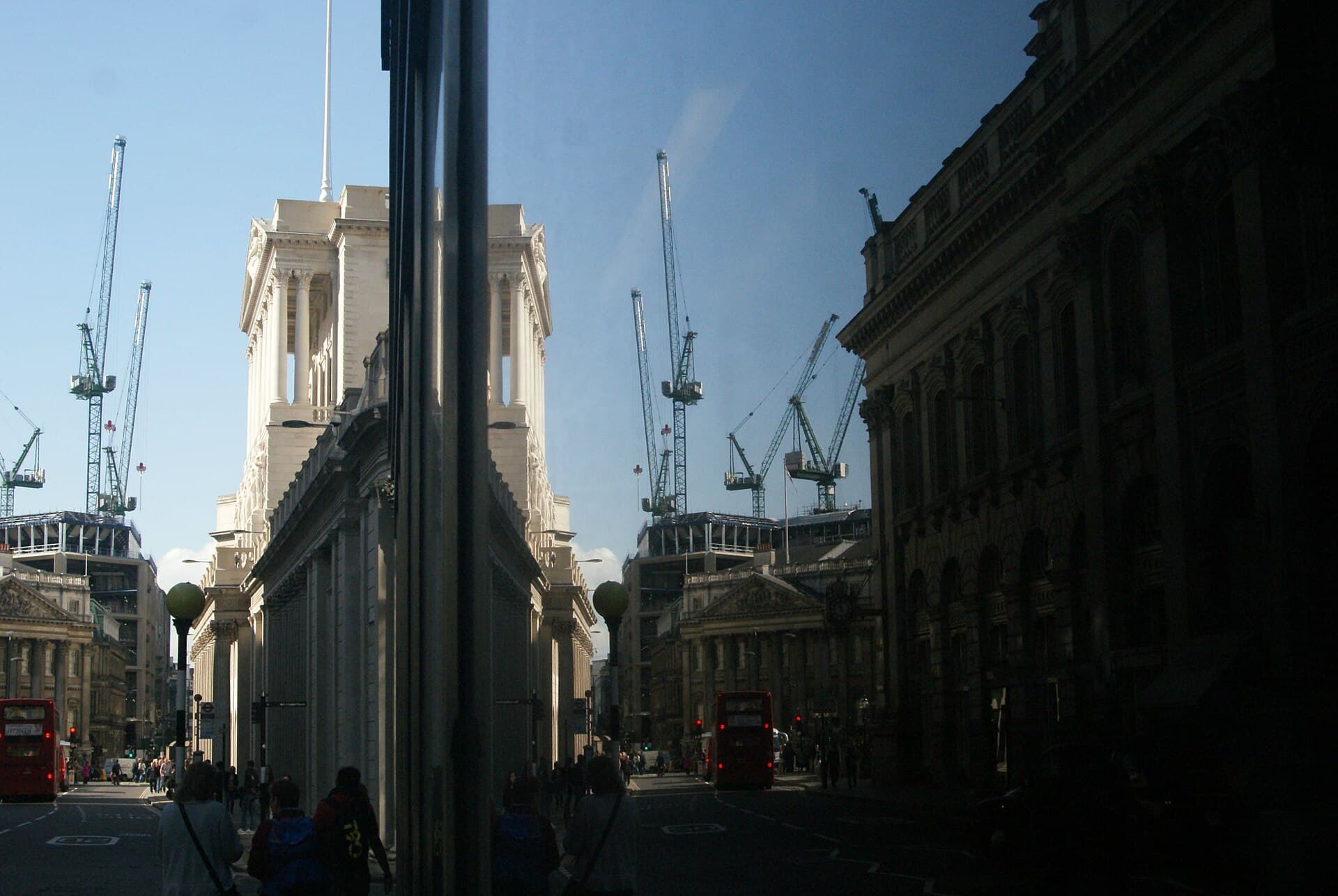 The Bank of England on Threadneedle Street in the City of London, reflected in a glass building with red buses and cranes in the background