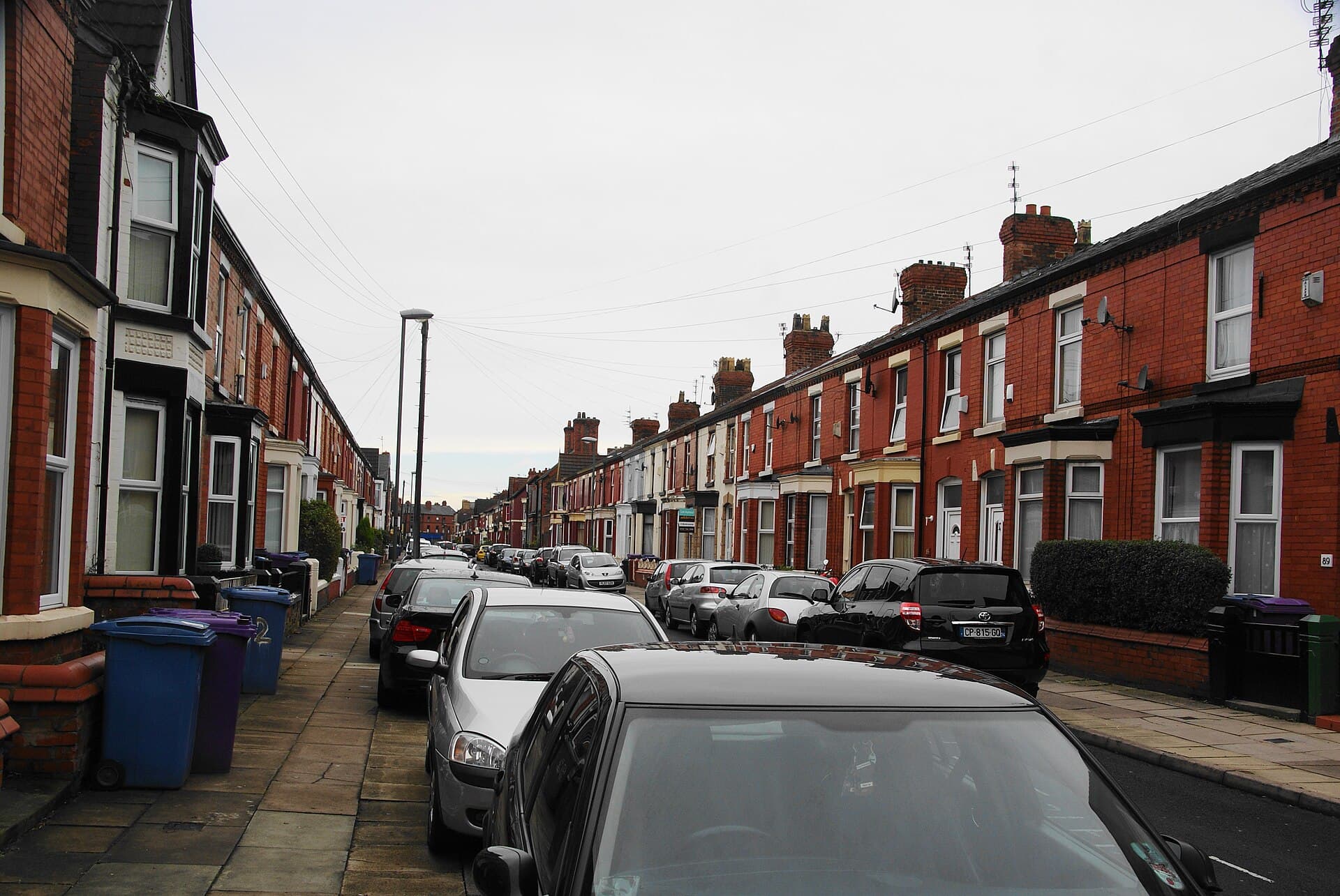 A long street of red-brick Victorian terraced houses with bay windows, wheelie bins and parked cars