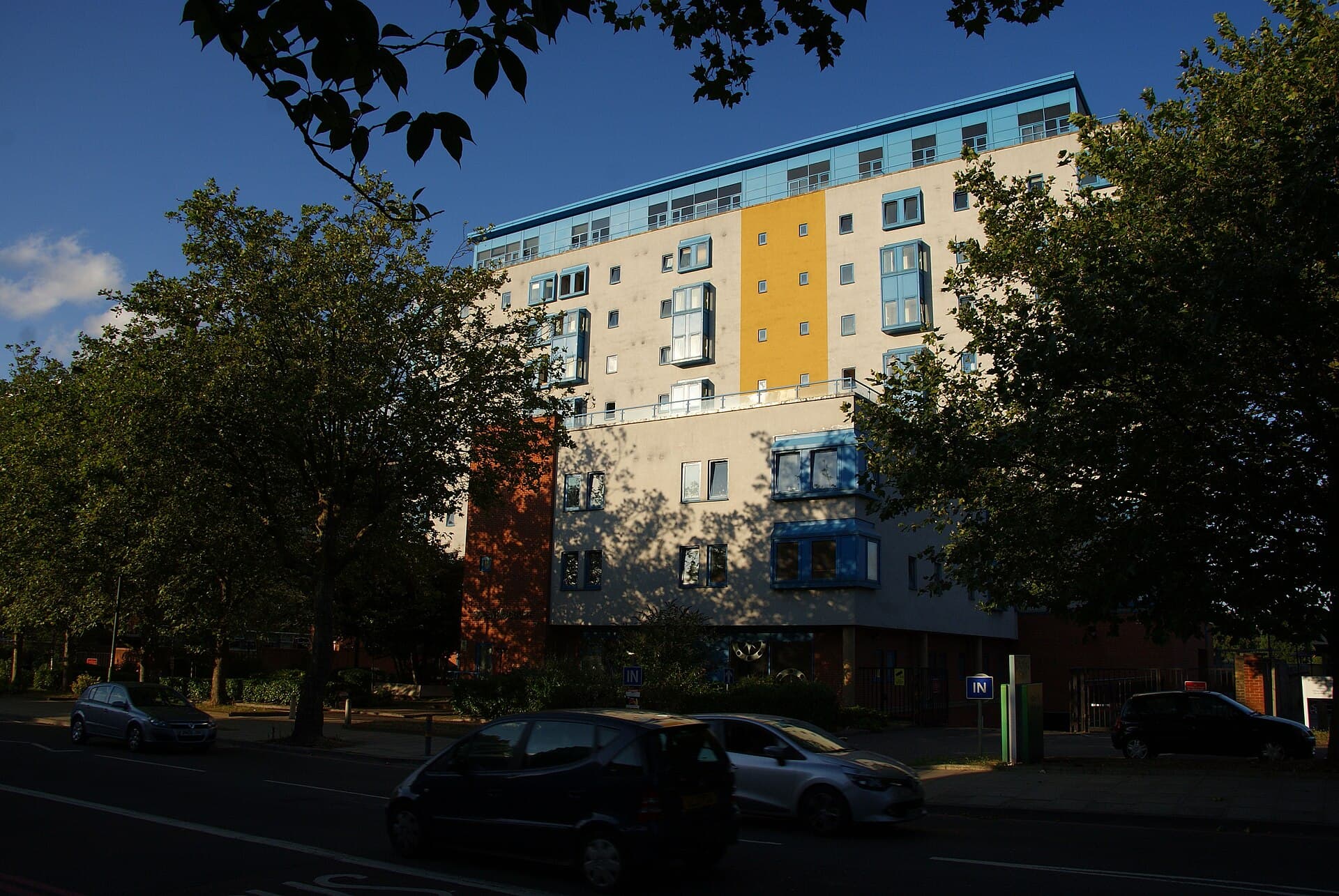A modern block of flats with blue and yellow panels behind trees on London Road in Norbury, south London, in evening sun