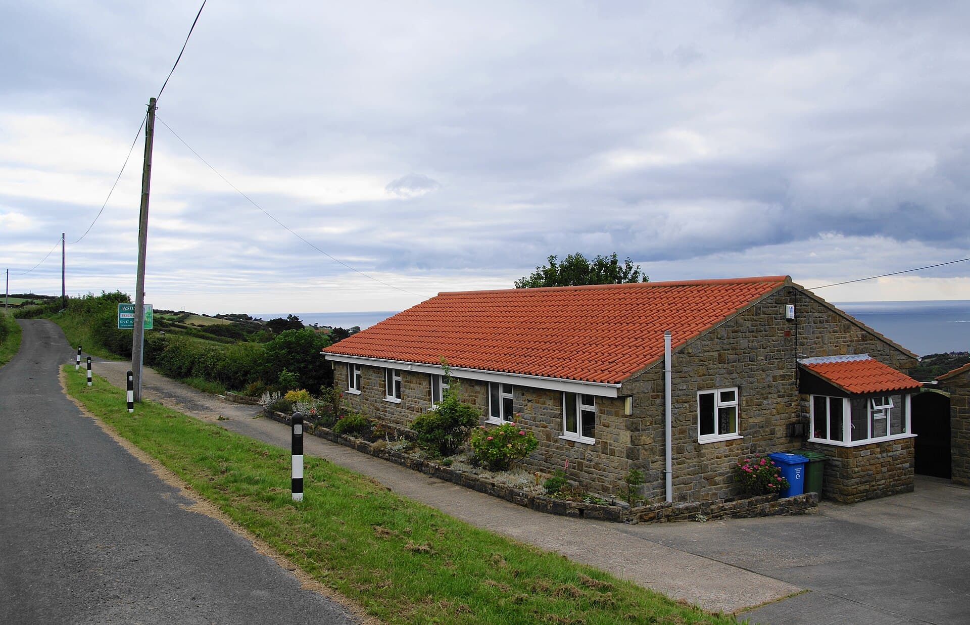 A stone bungalow with an estate agent's For Sale board on a country lane above the sea