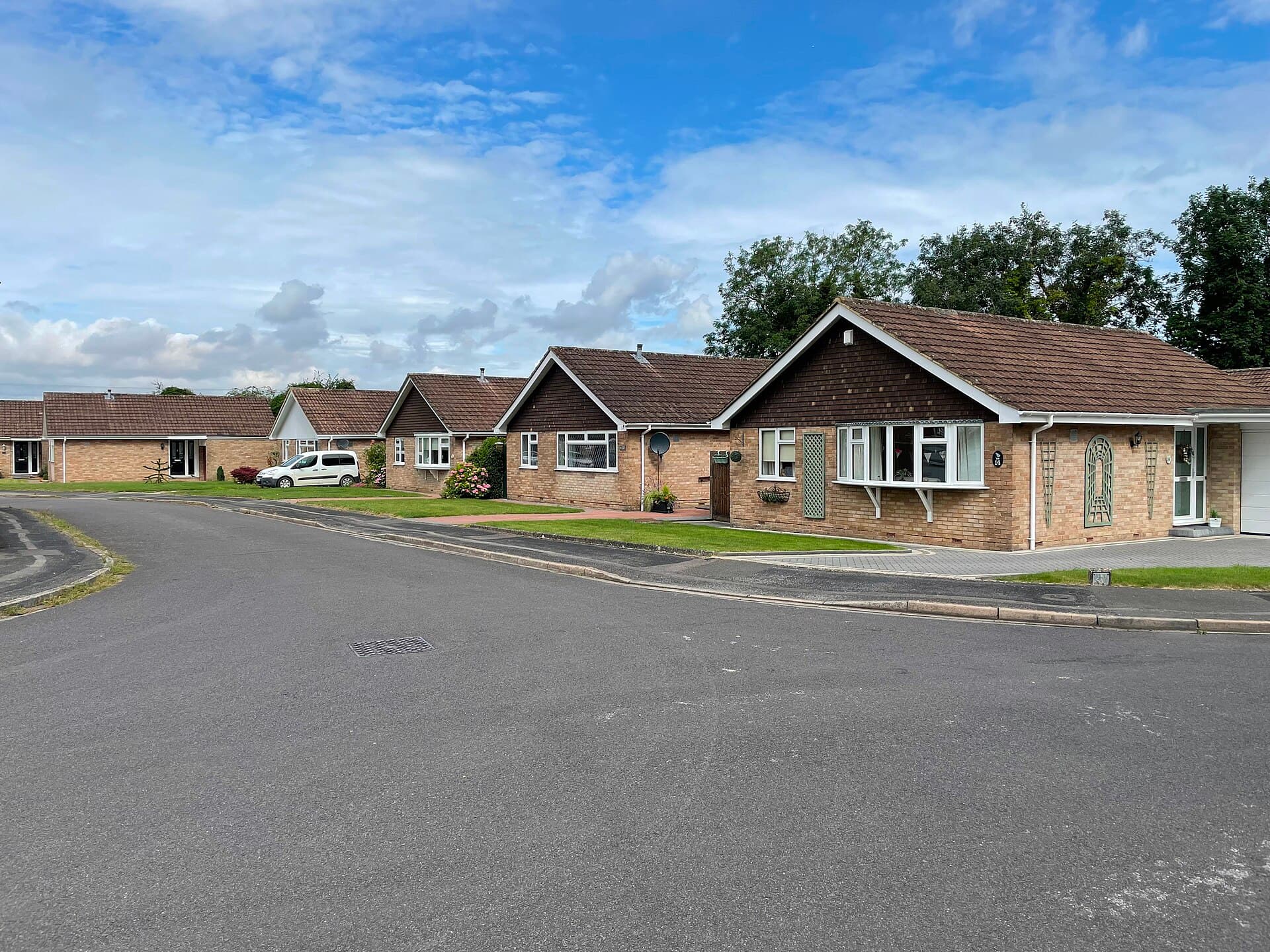 A quiet residential close of brick bungalows with neat front lawns and white bay windows under a bright, cloudy sky