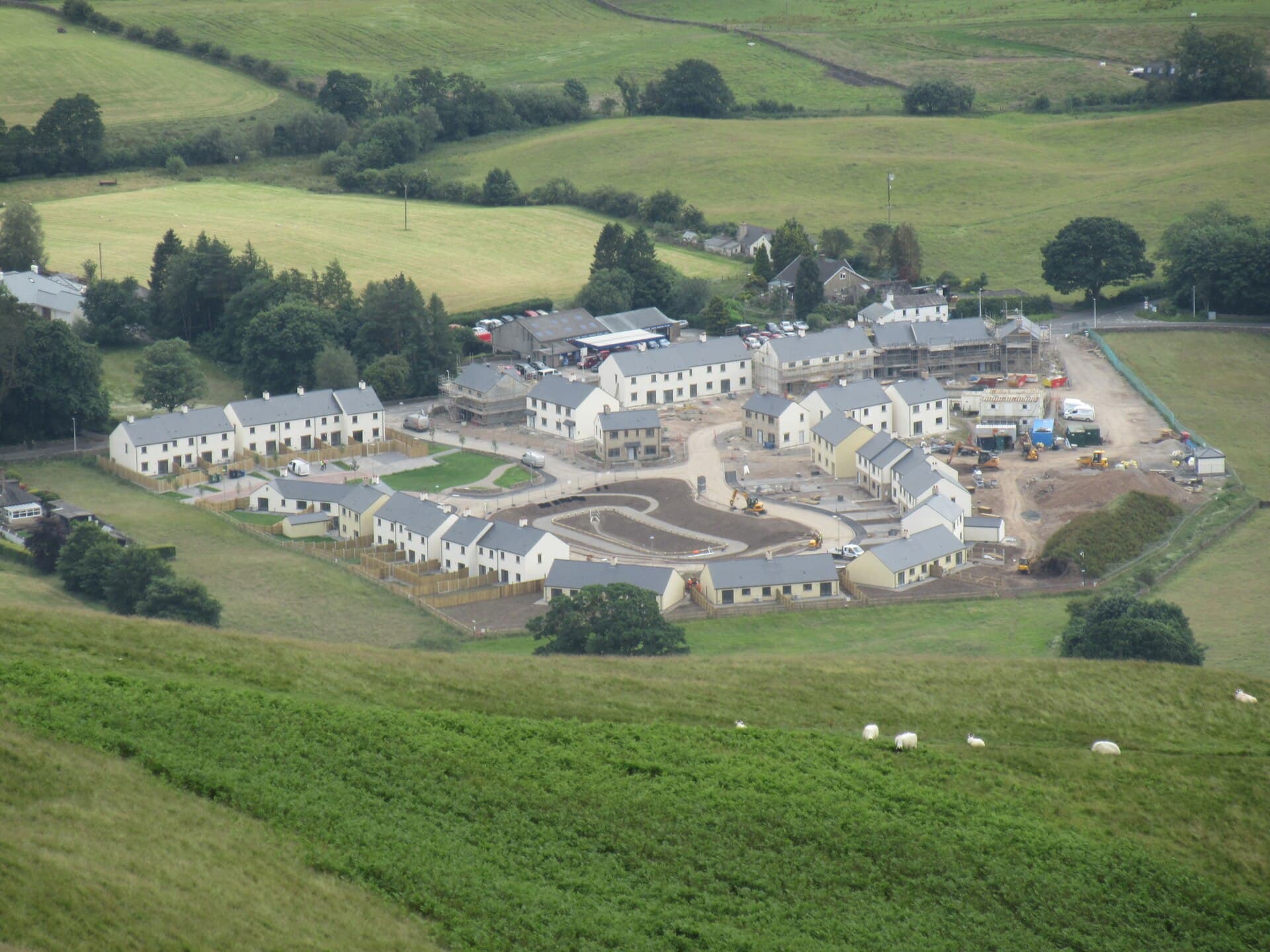 A new housing estate of terraced and semi-detached homes under construction in a green valley, seen from a hillside with sheep grazing