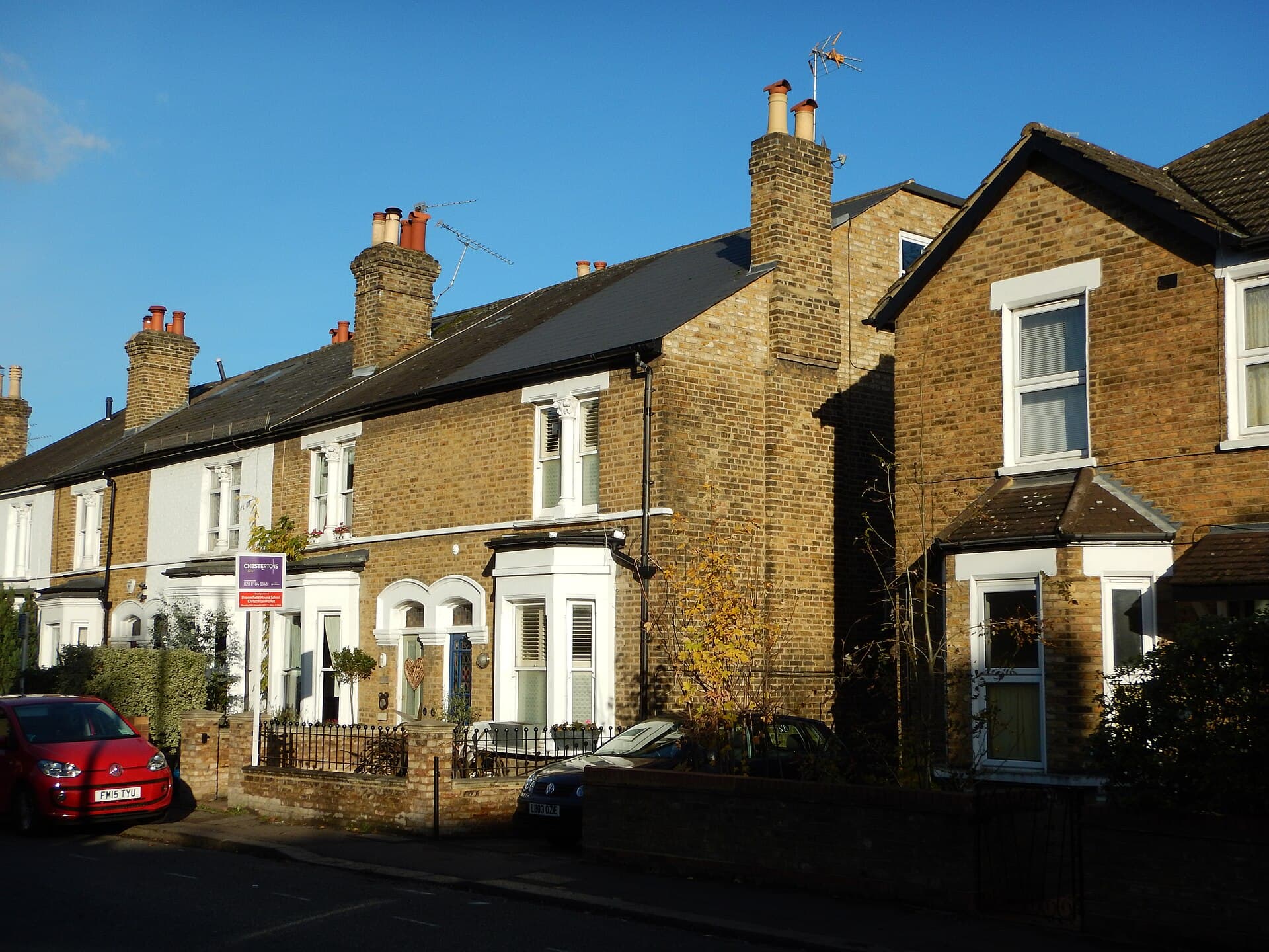 A row of brick Victorian terraced houses with white bay windows on a sunny residential road, with an estate agent's board outside one house and cars parked at the kerb