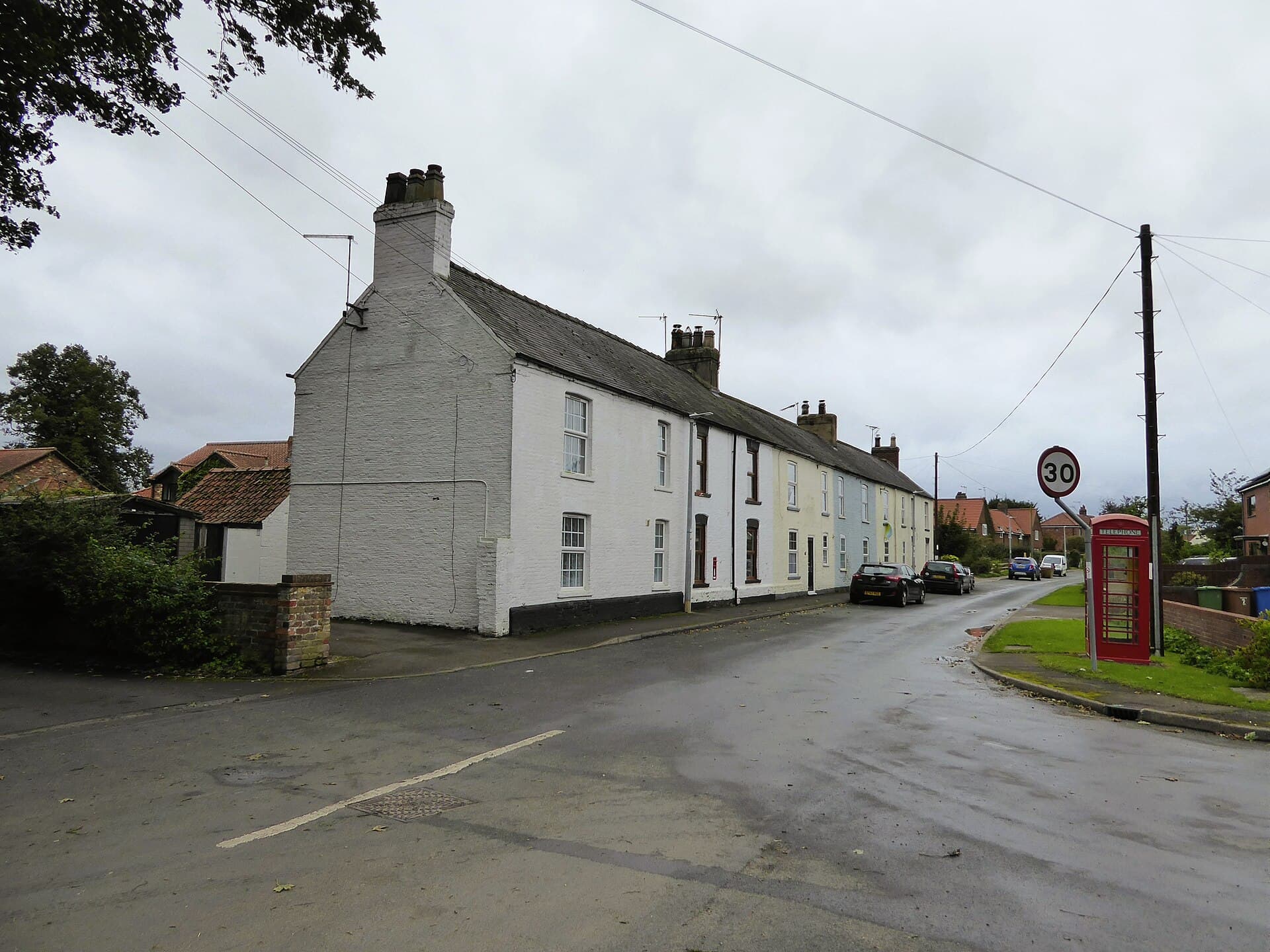 A row of painted terraced cottages on a wet village main street with parked cars, a red telephone box and a 30mph sign under a grey sky