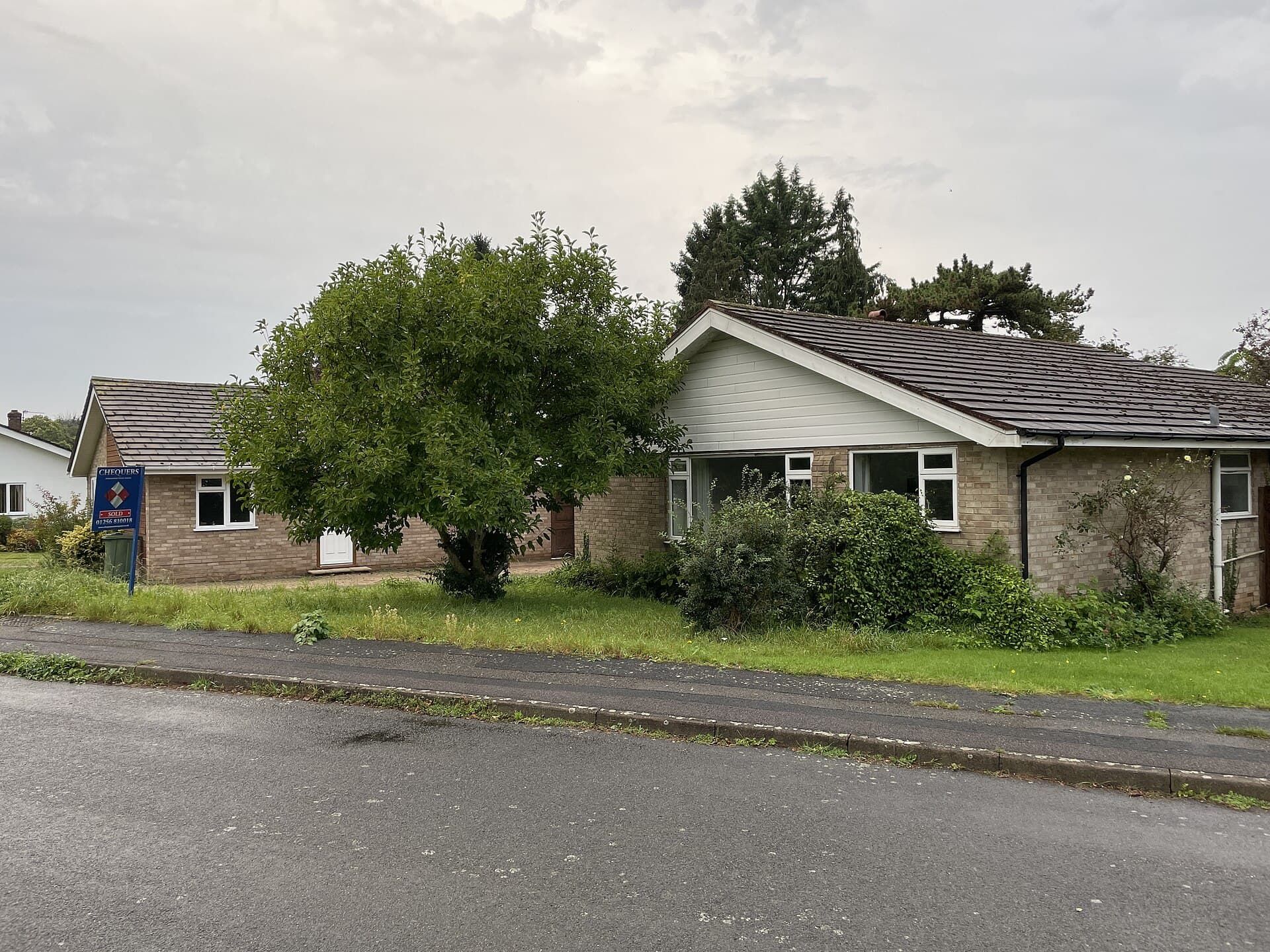 A brick bungalow with an overgrown front garden and an estate agent's board on the lawn, on a quiet residential road under a grey sky