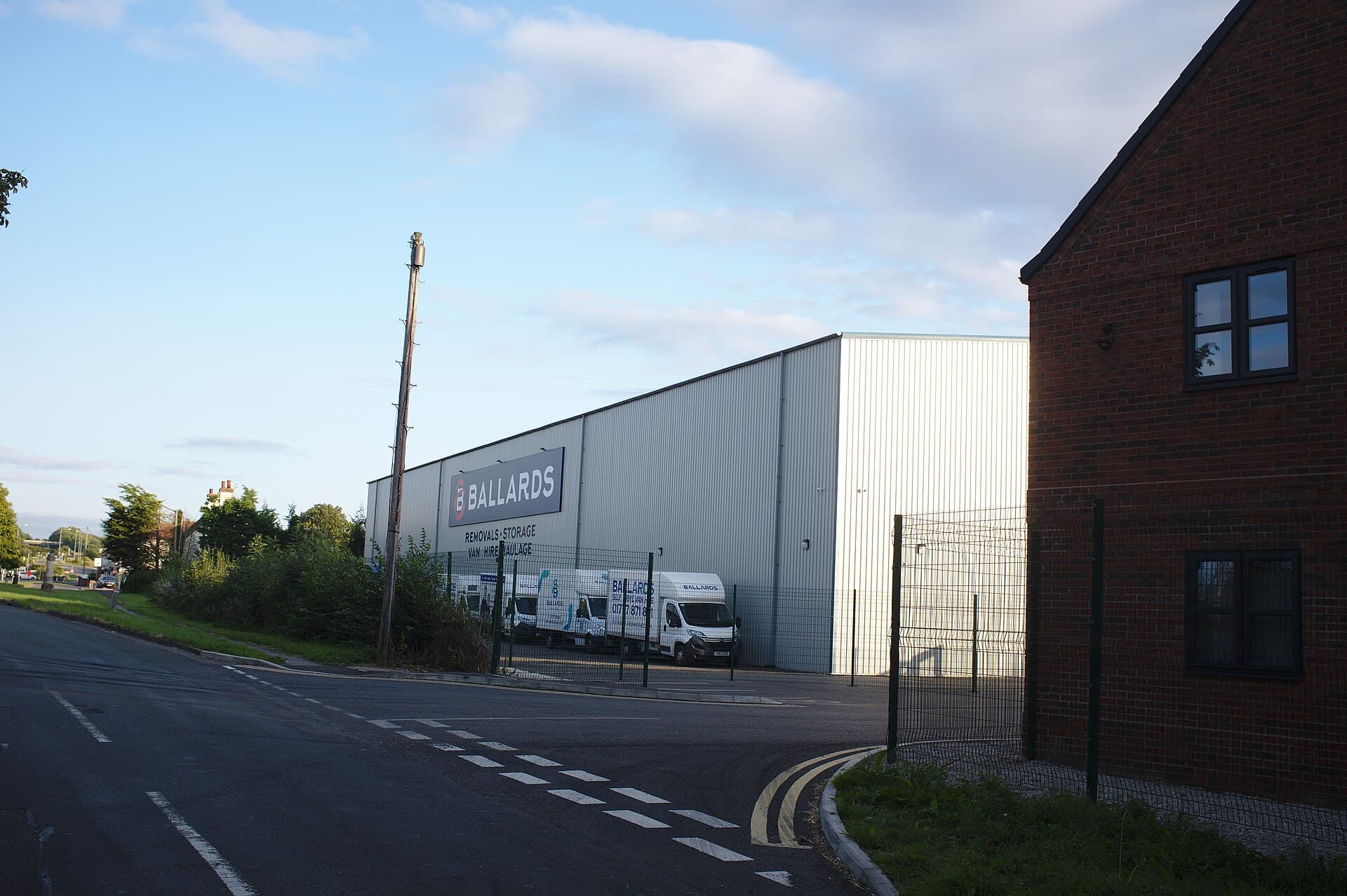 A removals and storage depot beside a road in evening light, with a row of white removal vans parked behind a fence