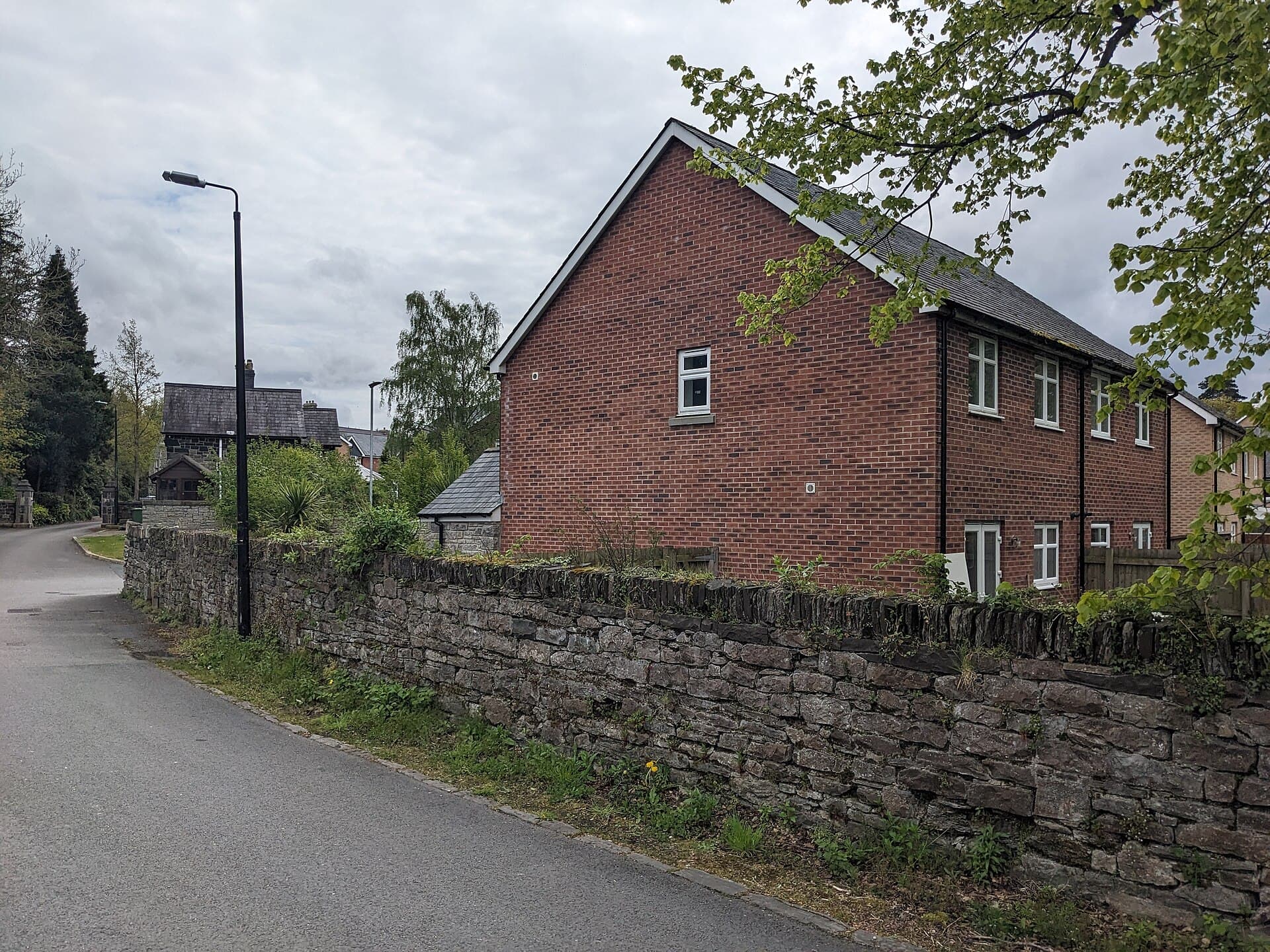 A red-brick modern house behind an old stone wall on a quiet residential lane with a street lamp and trees