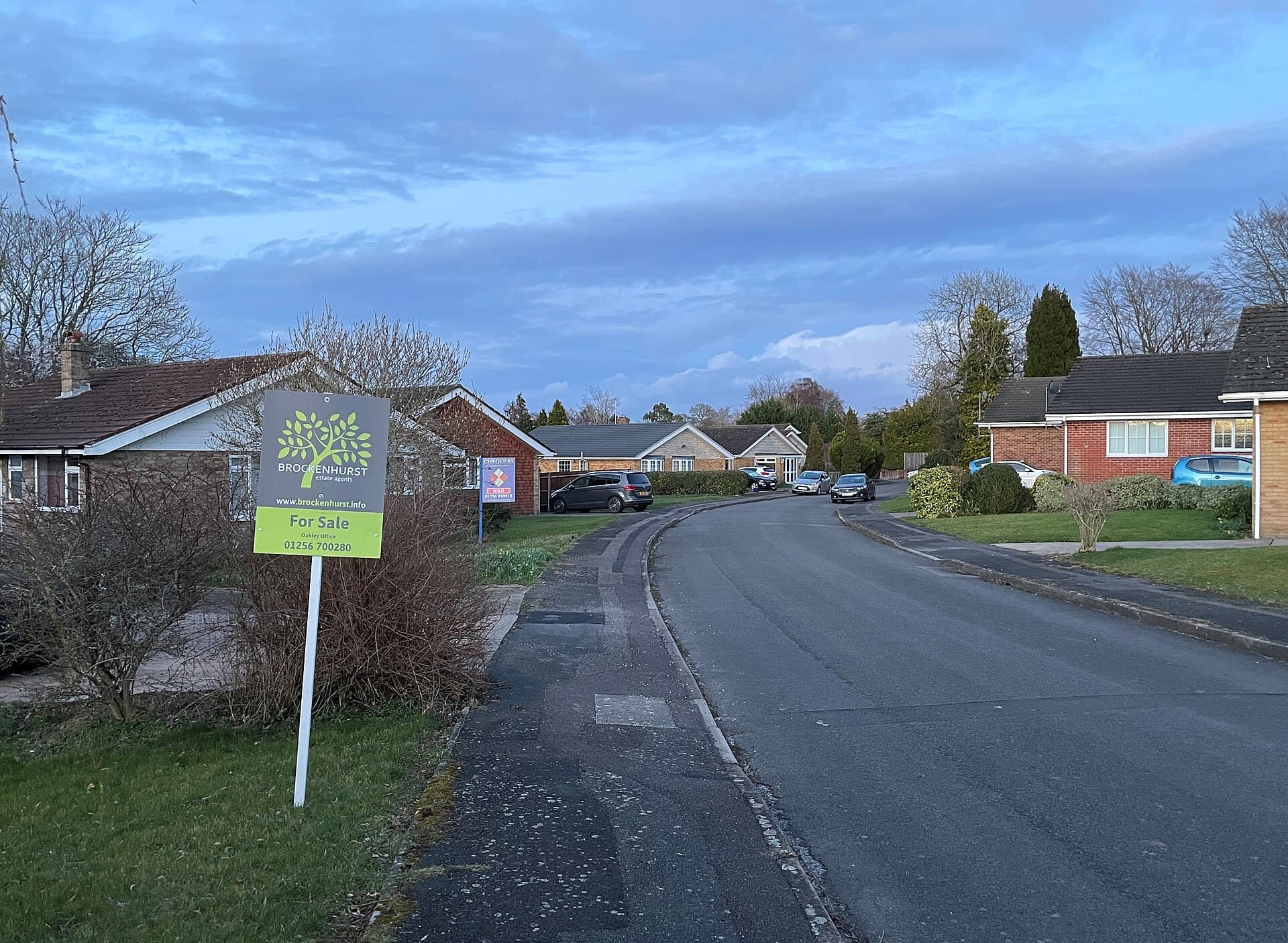 A quiet suburban road of bungalows at dusk with an estate agent's For Sale board in the foreground and a second board further along the street